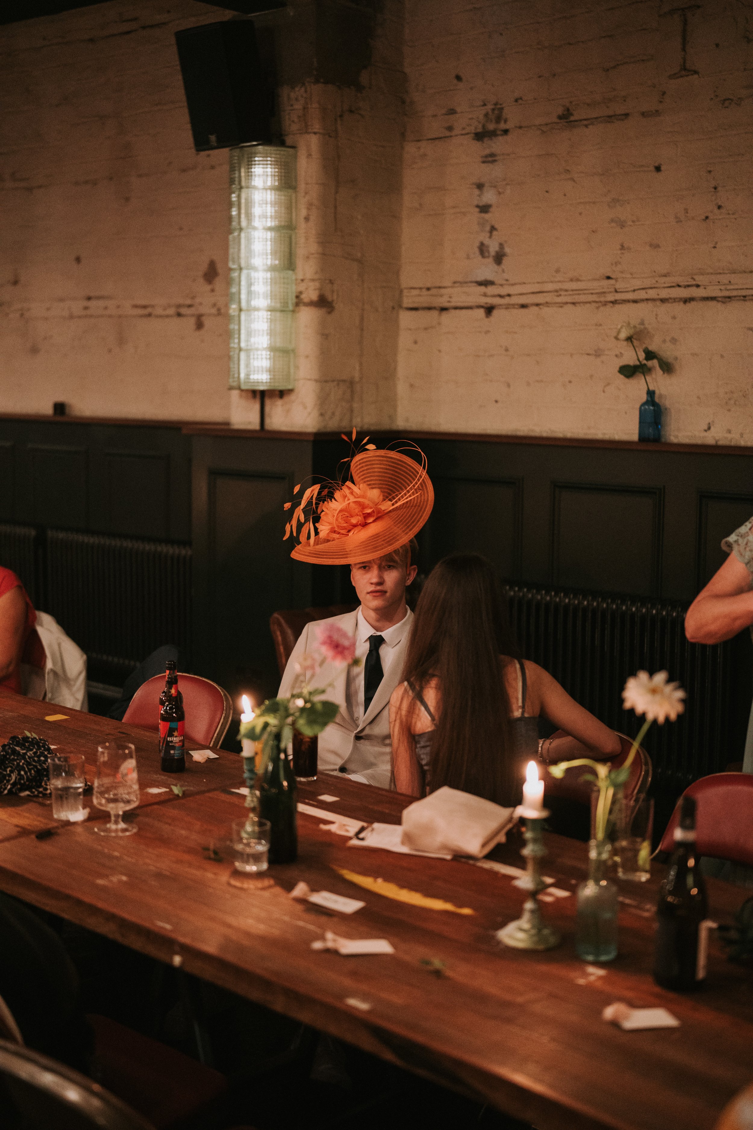 Person wearing an orange fascinator sitting at a wooden table with flowers and candles in a rustic setting.