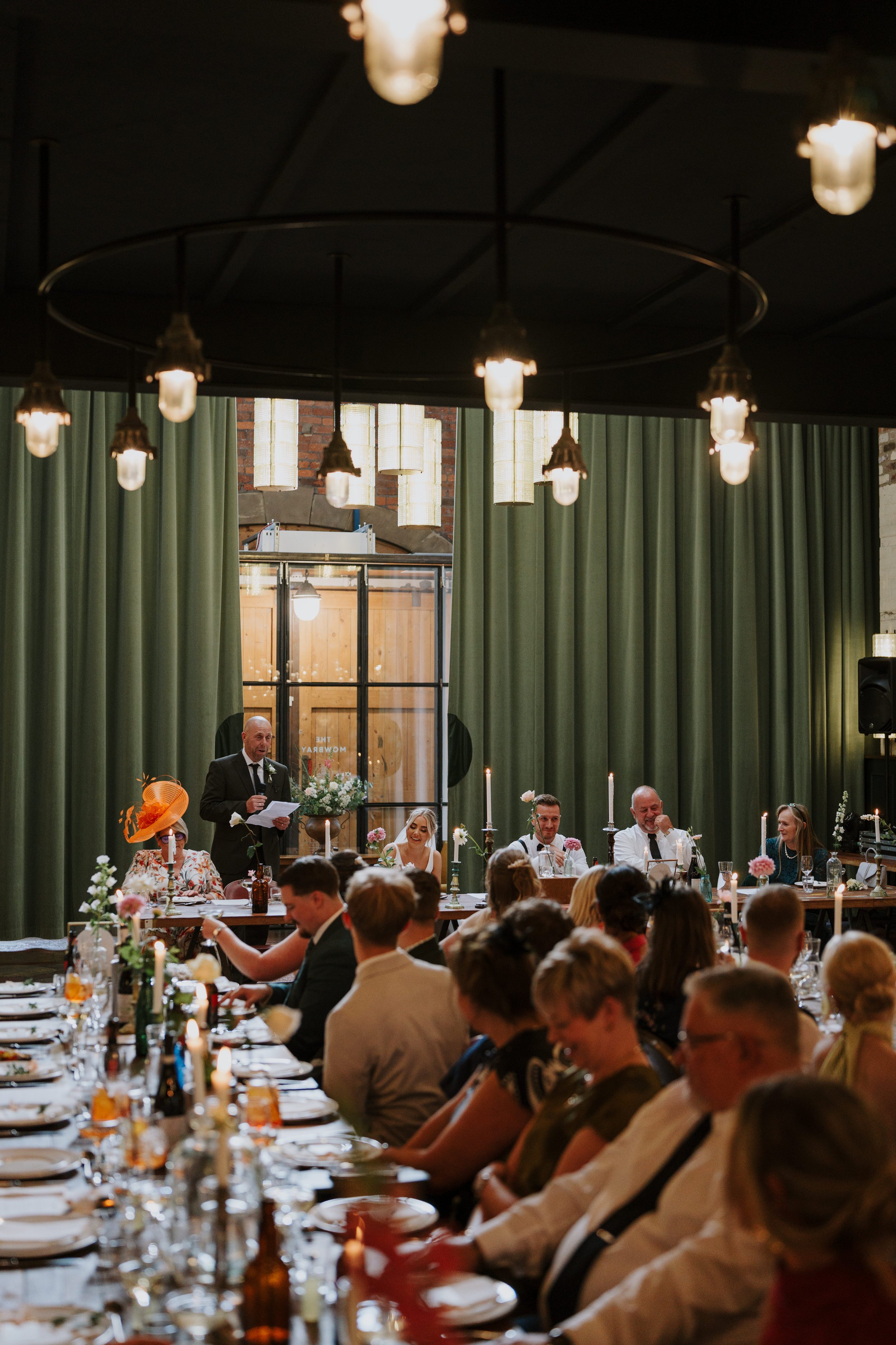 Wedding reception with guests seated at tables, a speaker giving a speech in front of green curtains, and decorative lighting overhead.