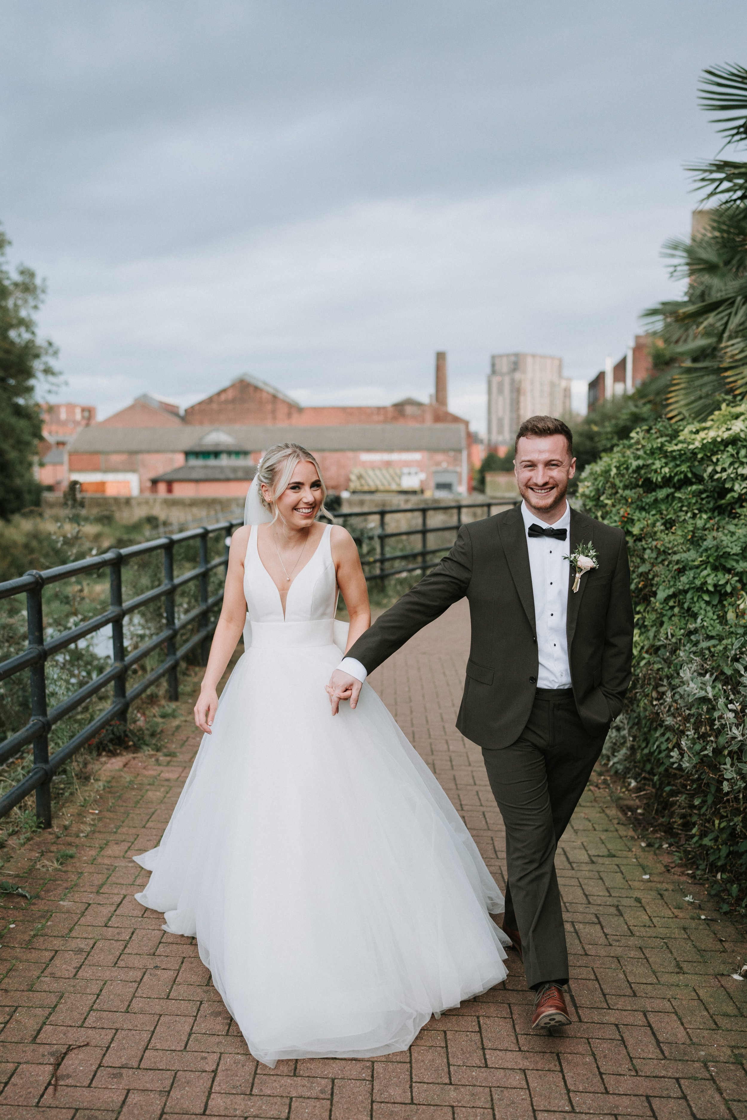 Bride and groom walking hand in hand outdoors, smiling, with industrial buildings in the background.