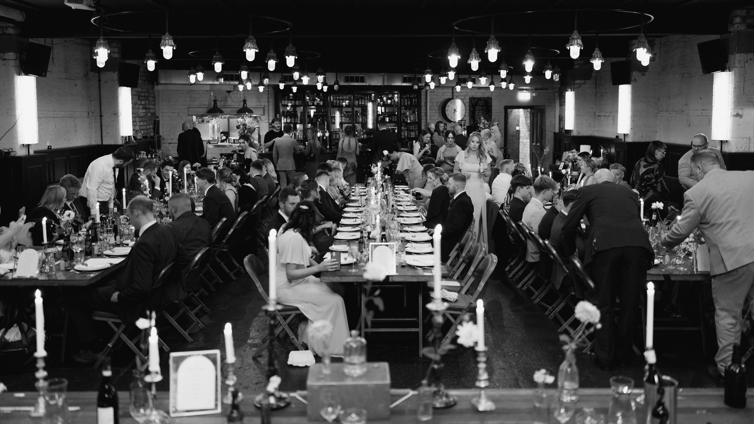 Black and white photo of a wedding reception with guests seated at long tables adorned with candles.