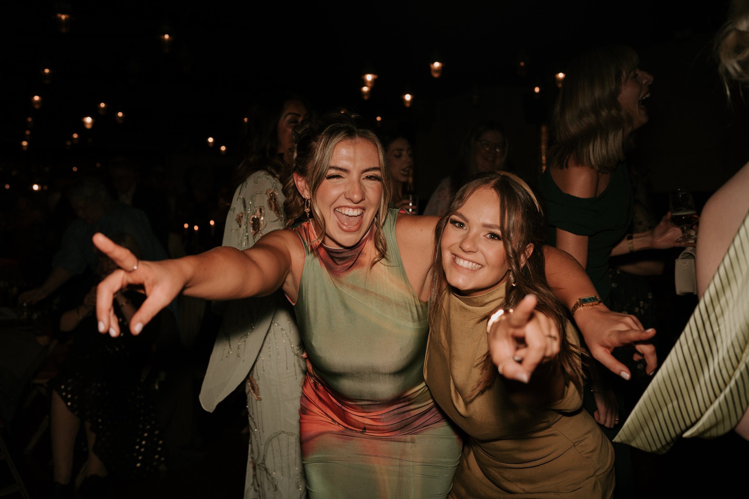 Two women smiling and pointing at the camera in a dimly lit party setting, with blurred people and lights in the background.