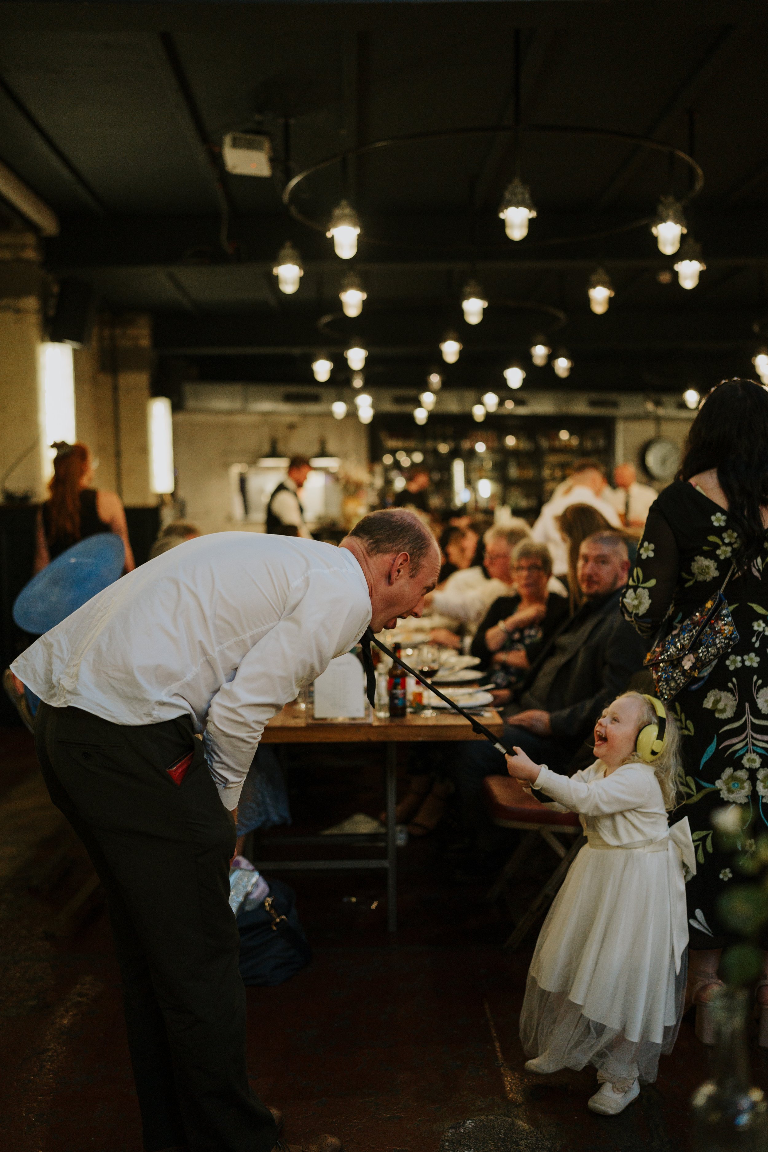 Man playfully interacting with smiling child wearing earmuffs at a dinner event.