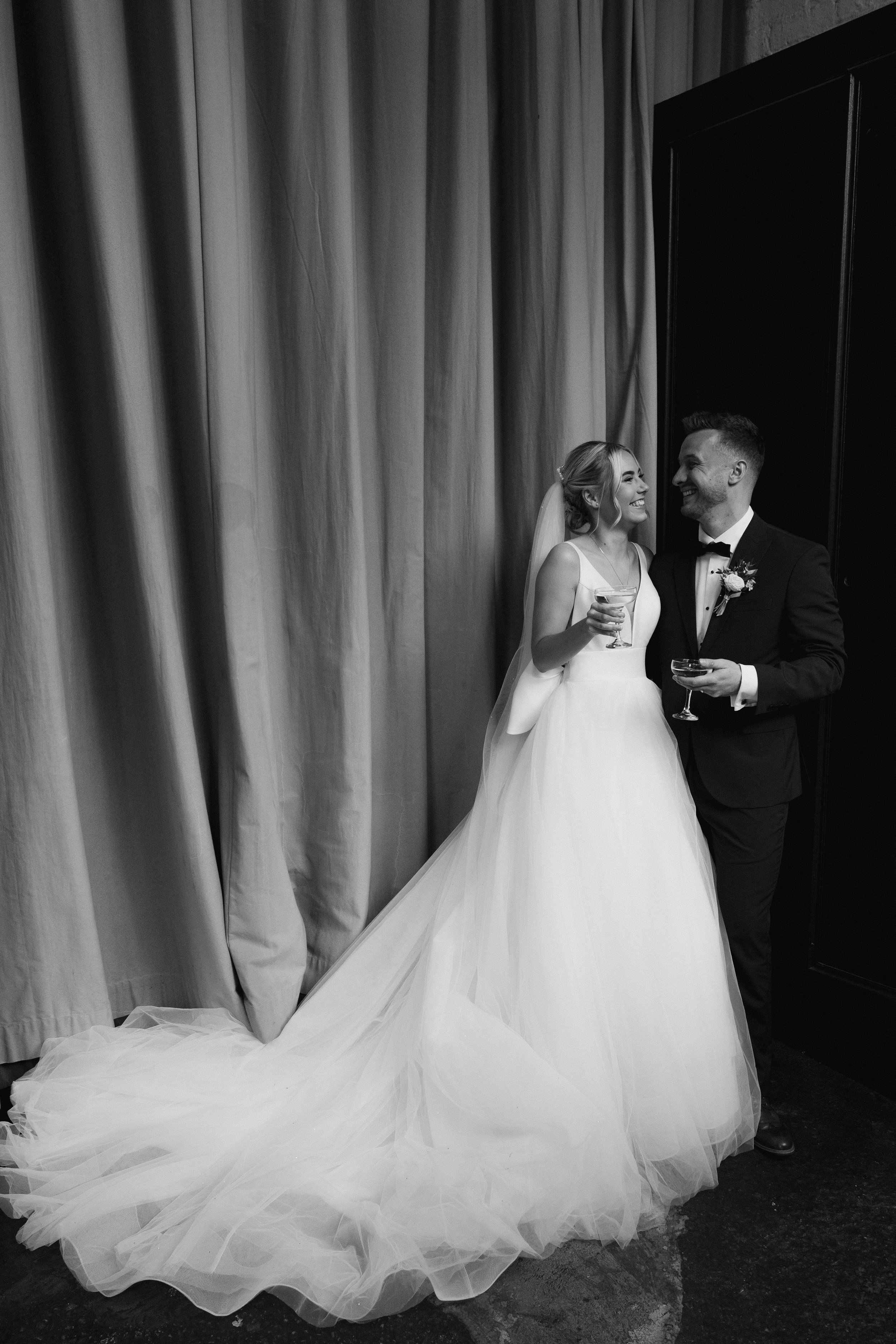 Black and white photo of a bride and groom smiling, dressed in wedding attire, holding drinks, standing near a curtain.