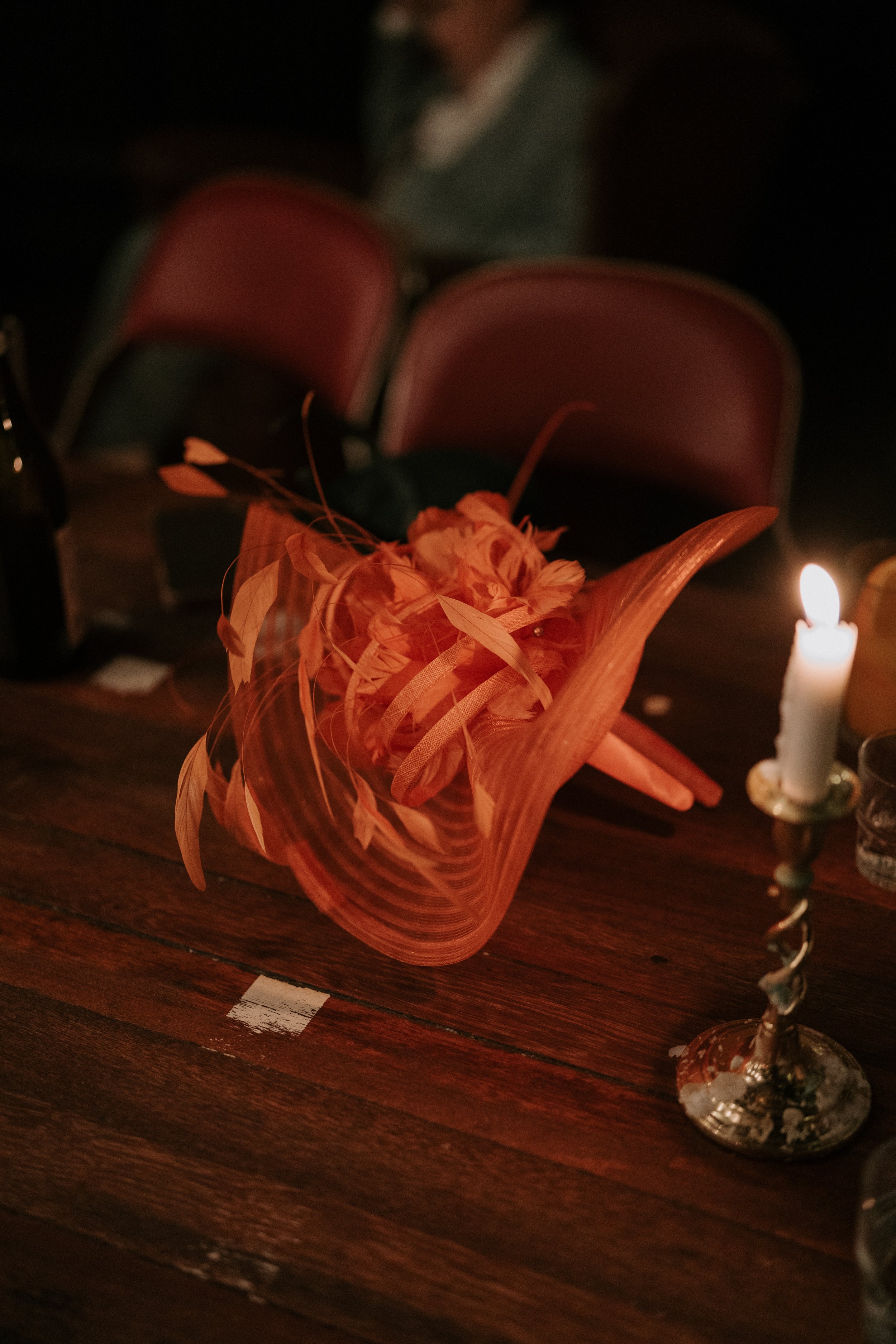 Decorative orange hat with feathers on wooden table beside a lit candle in holder, bottles, and glasses, with red chairs in the background.