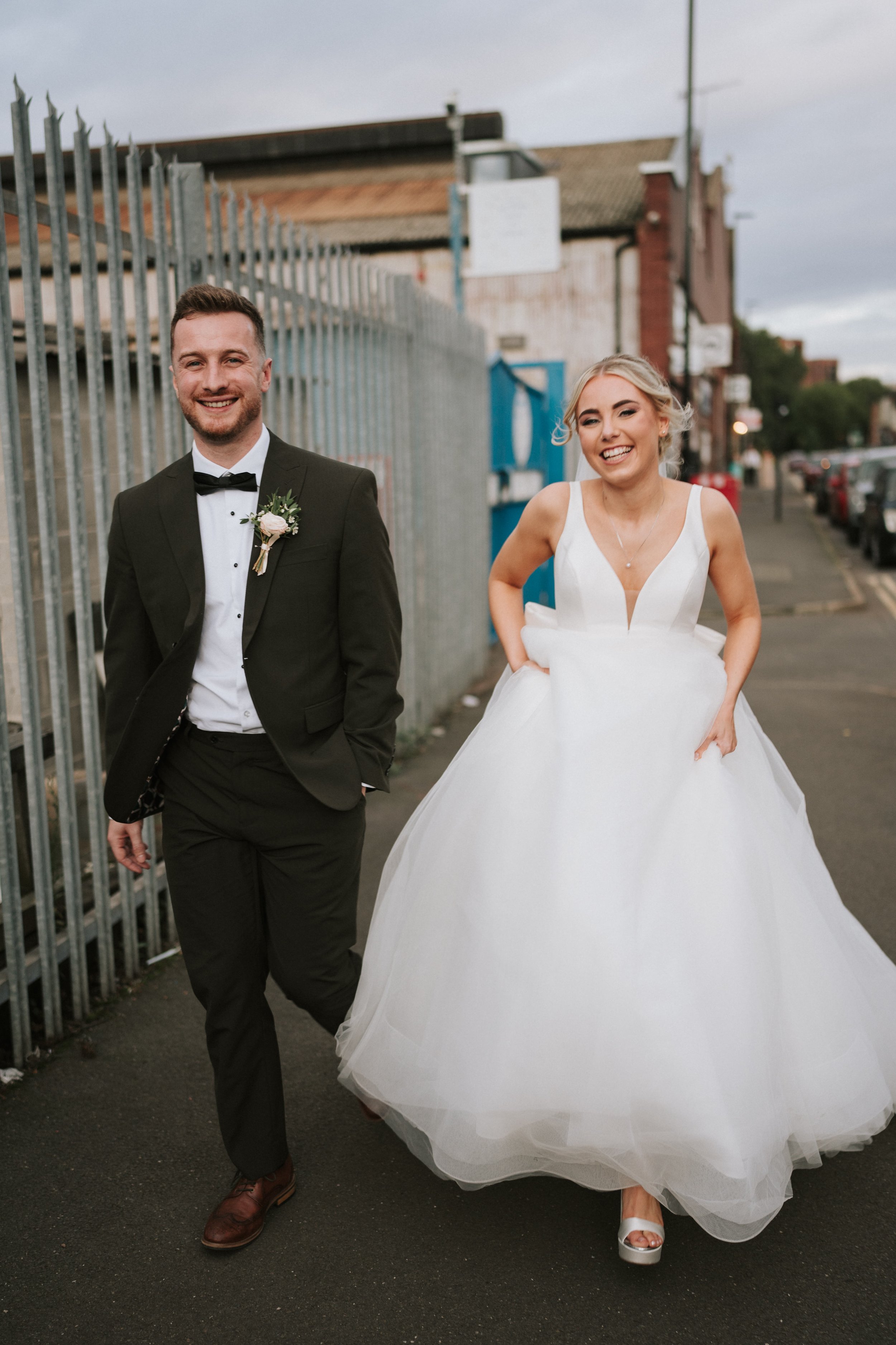 Bride and groom walking outside, bride in white gown, groom in black tuxedo, smiling.