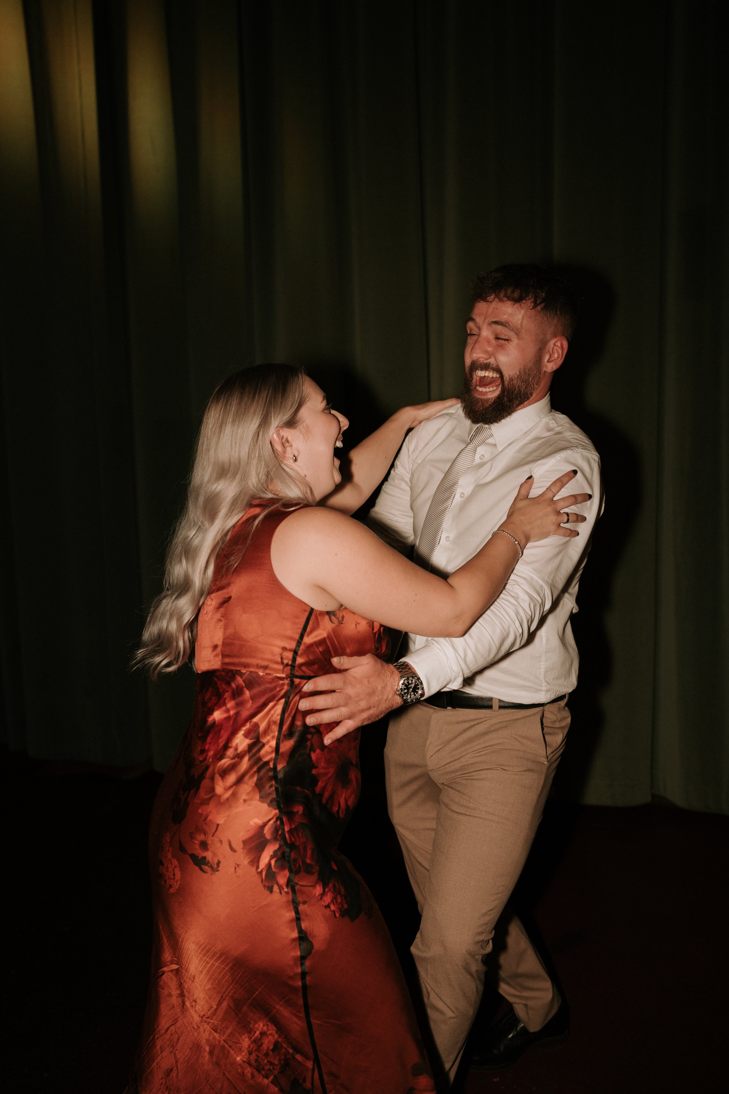 A man and woman dancing together, smiling and dressed in semi-formal attire. The woman wears a floral dress and the man wears a white shirt and tie.