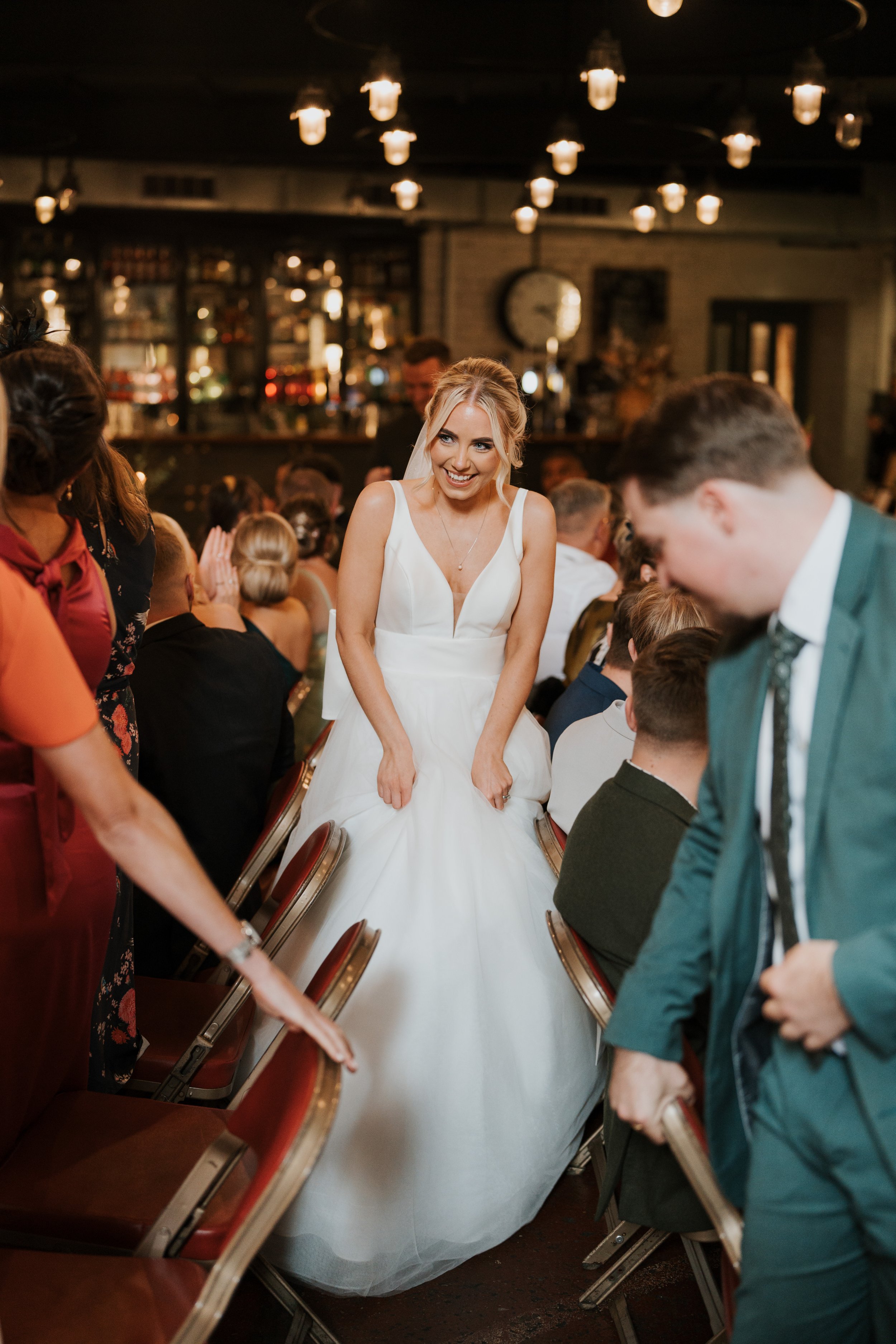 Bride in white gown smiling and navigating between seated guests indoors, decorative lights overhead.
