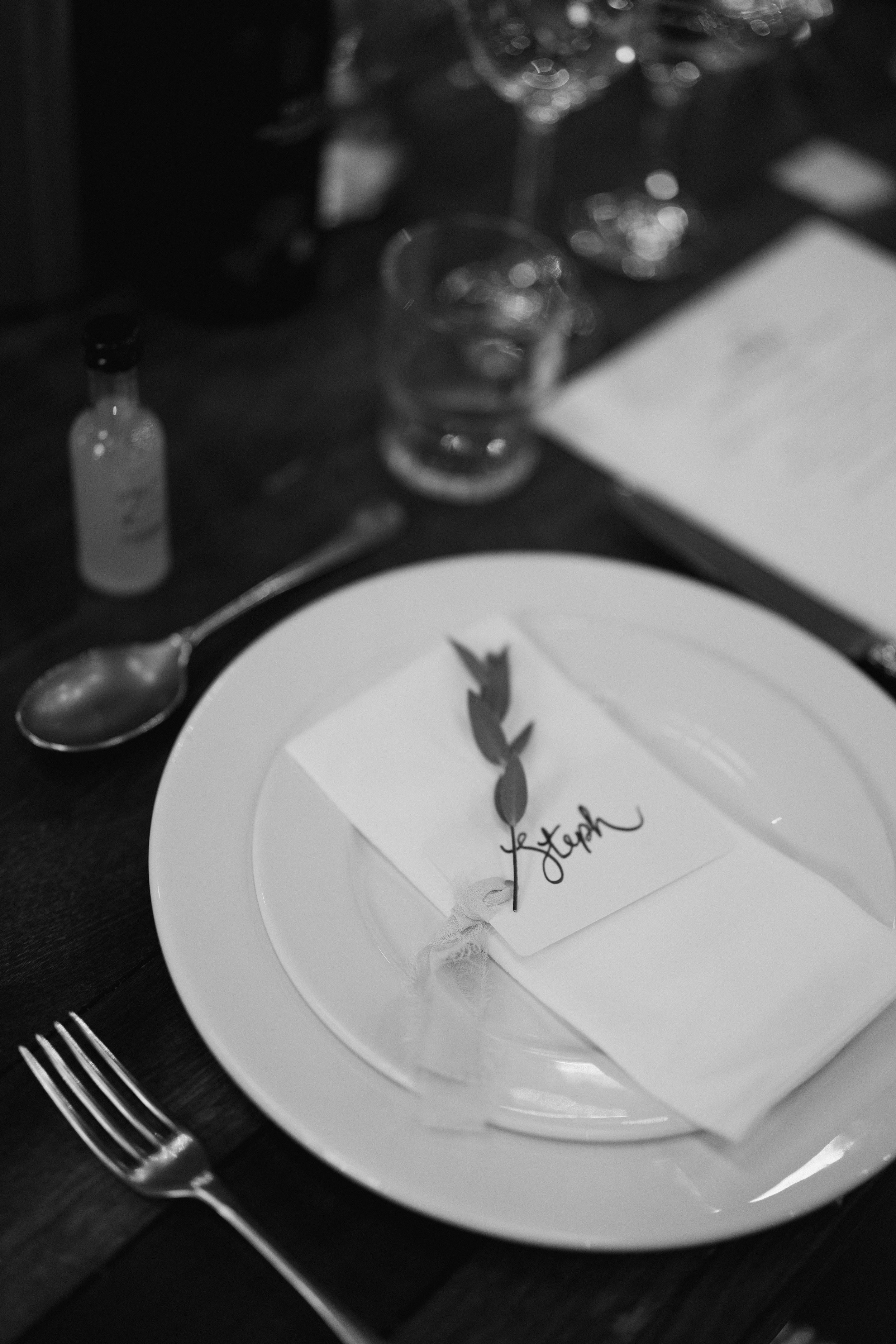 Black and white photo of an elegant dinner table setting with a plate, napkin, and name card with a leaf on top, surrounded by cutlery, glassware, and a small bottle.