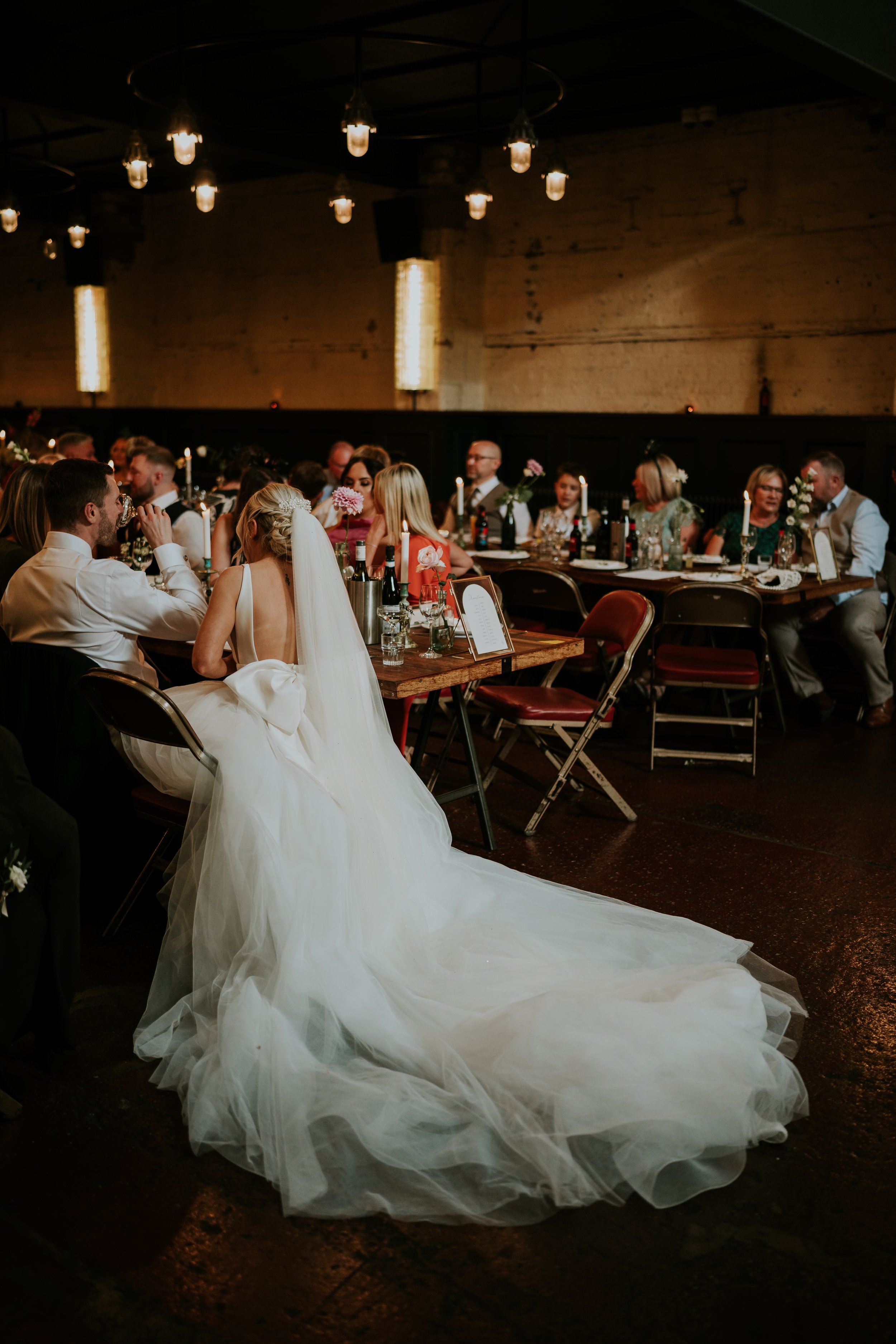 Wedding reception scene with a bride in a long white dress sitting at a table surrounded by guests in a dimly lit room.