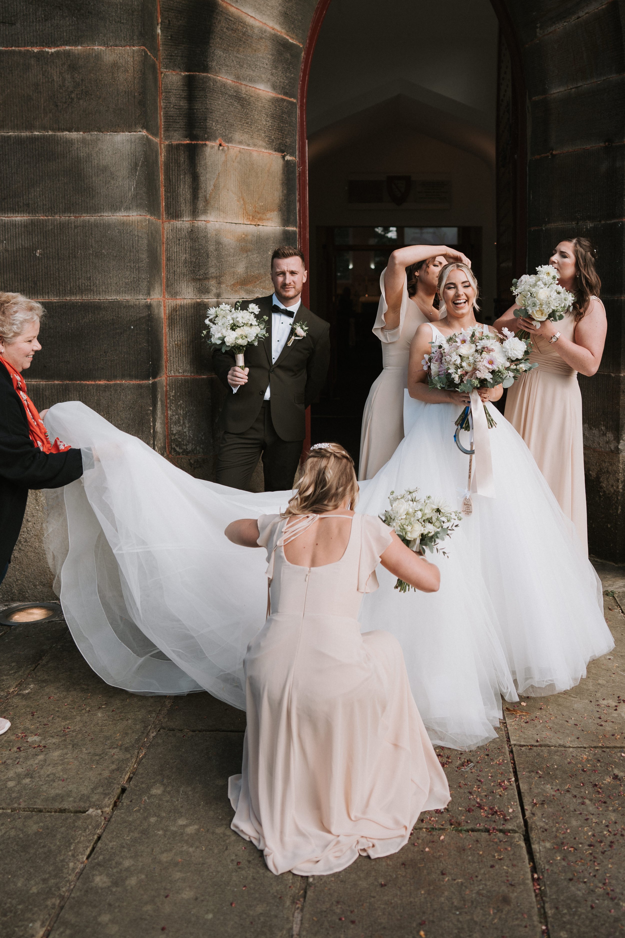 Wedding party with bride and bridesmaids holding bouquets, adjusting dress train, smiling outdoors.