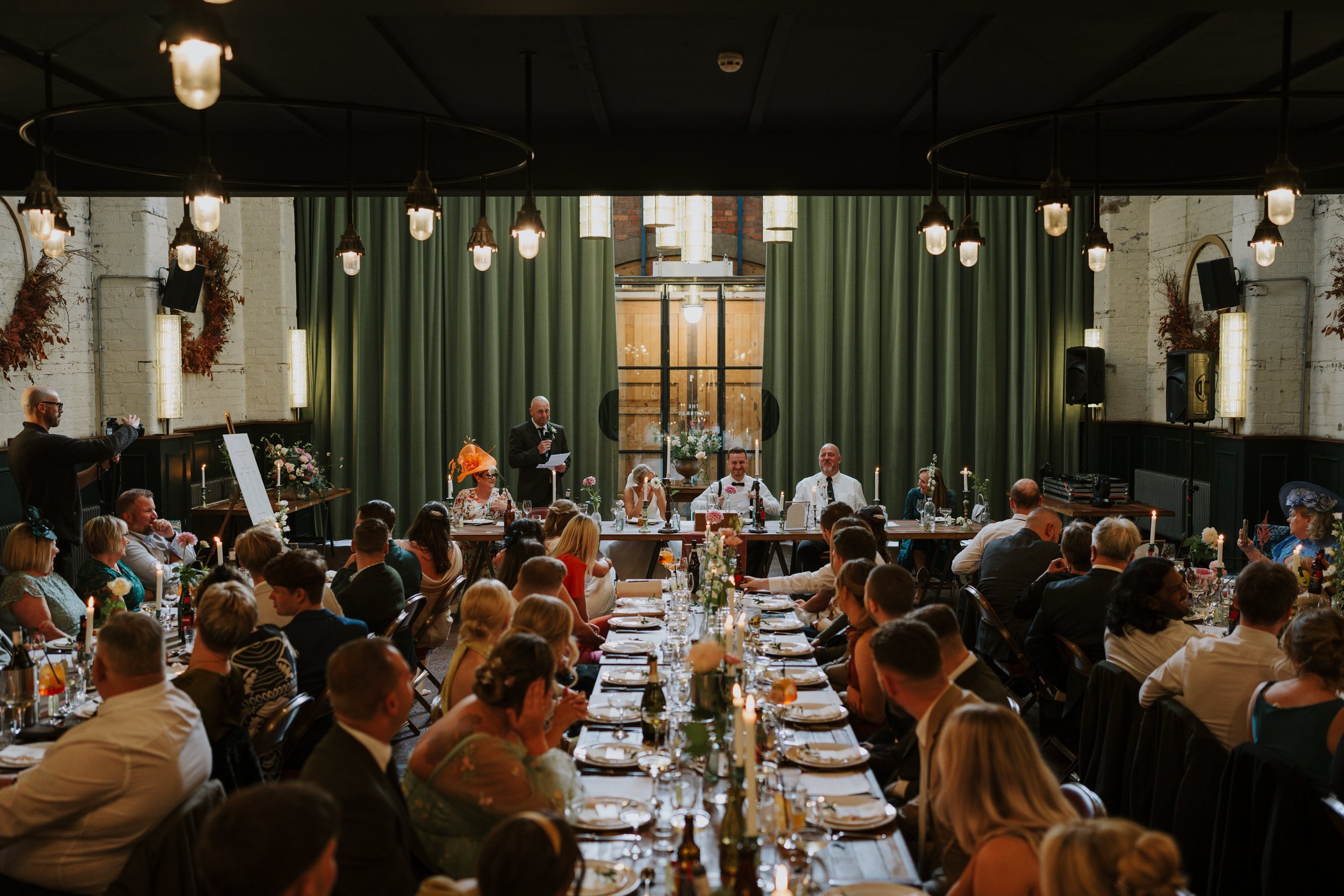 Wedding reception with guests seated at long tables, person giving speech at head table, elegant decor, dim lighting.