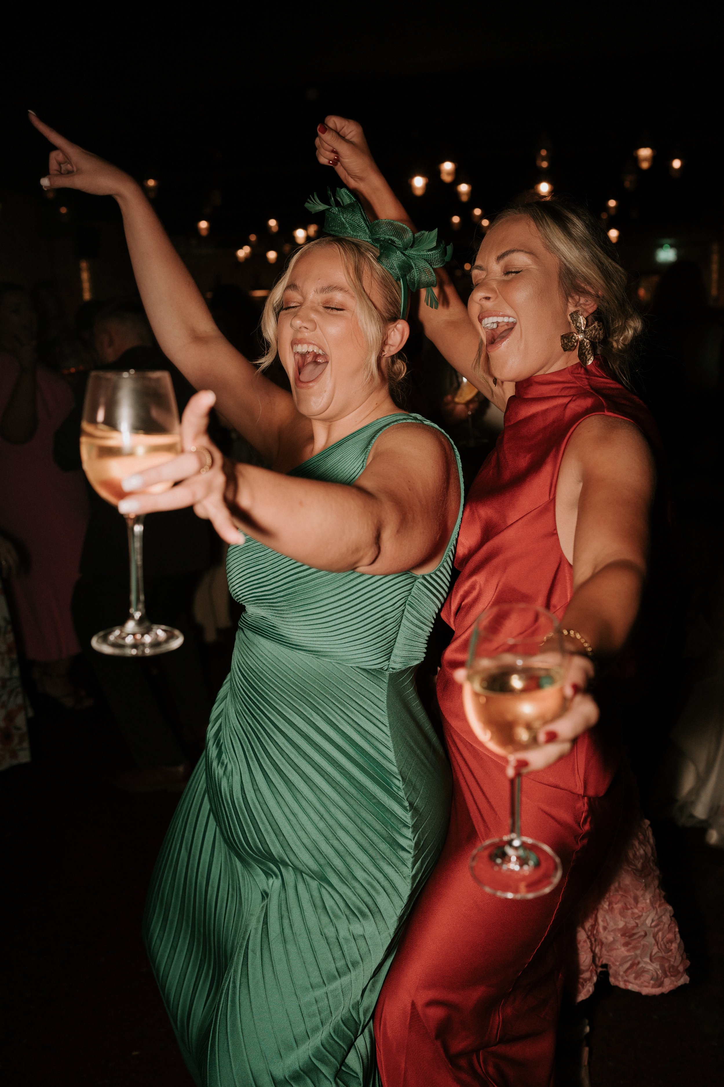 Two women dancing and celebrating at a party, holding wine glasses and dressed in vibrant green and red outfits.