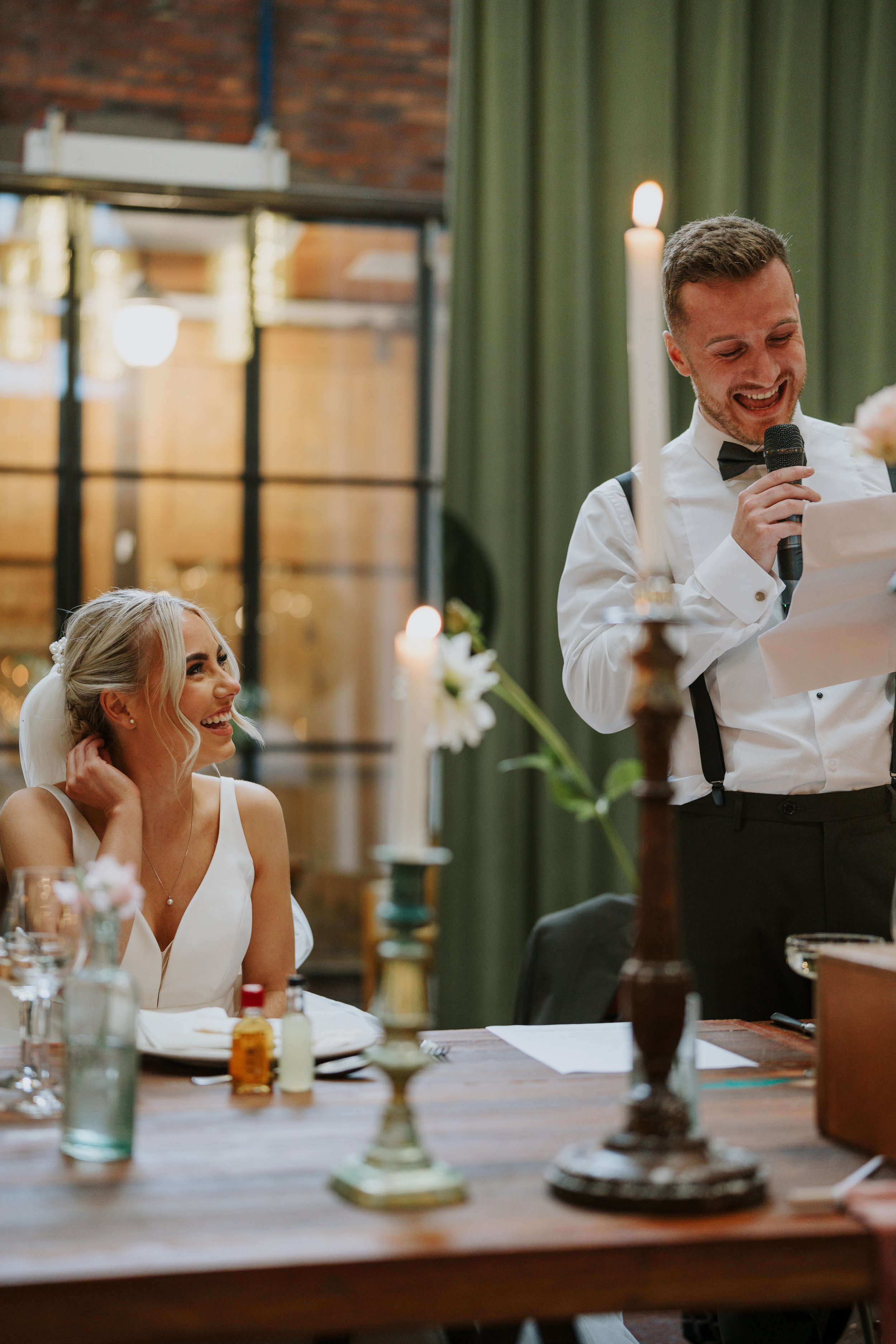 Bride smiling and groom giving speech at wedding reception.