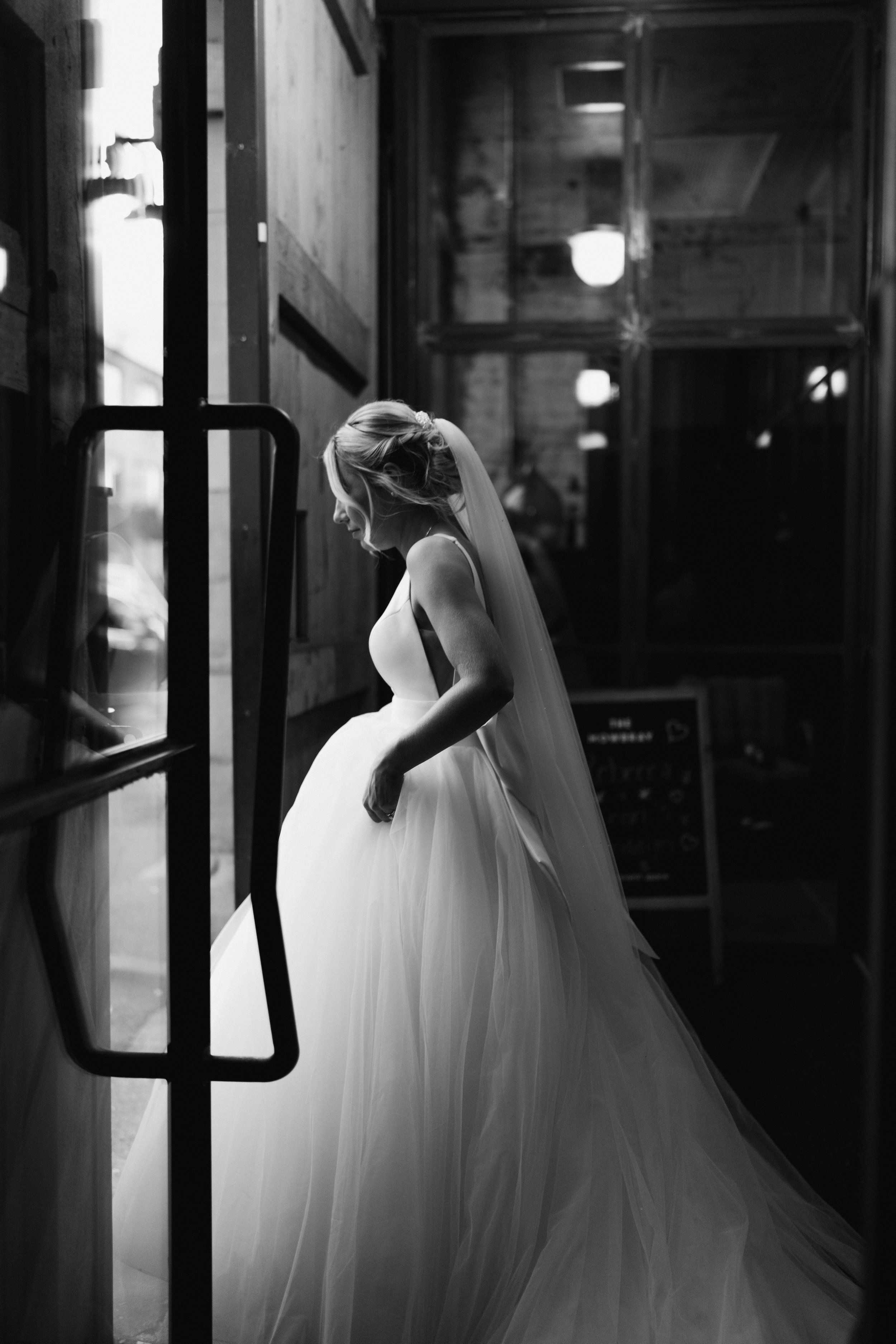 Black and white photo of a bride in a wedding dress near a glass door inside a venue.