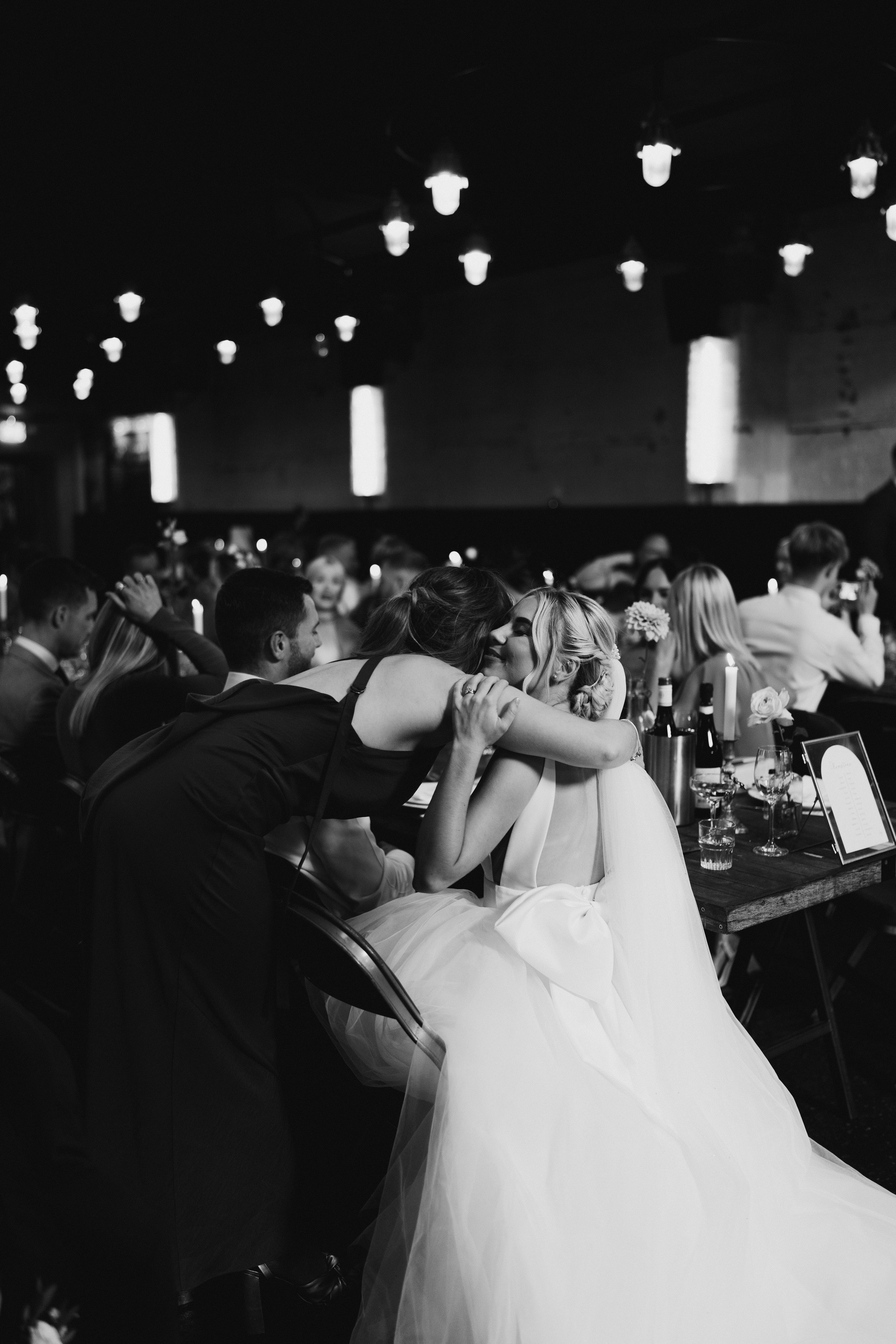 Black and white photo of two women hugging at a wedding reception, one in a bridal gown, with guests seated at tables in the background.