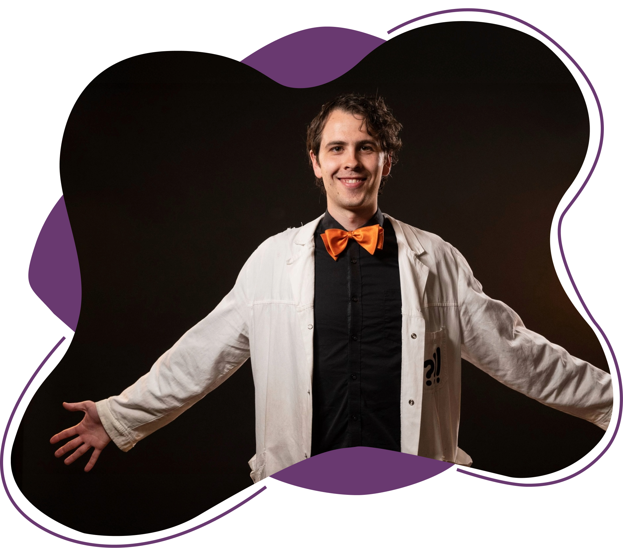 En ung mann smiler, iført en svart skjorte med oransje sløyfe, og en hvit labfrakk, med armene utstrukket i en åpen positur mot en svart bakgrunn.