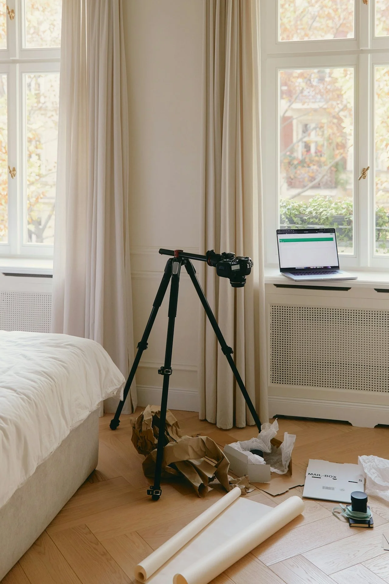 Camera on tripod, open laptop on windowsill, crumpled packing paper, mail box box, rolls of paper, and other miscellaneous items on hardwood floor next to bed in a bright room with large windows and cream curtains.