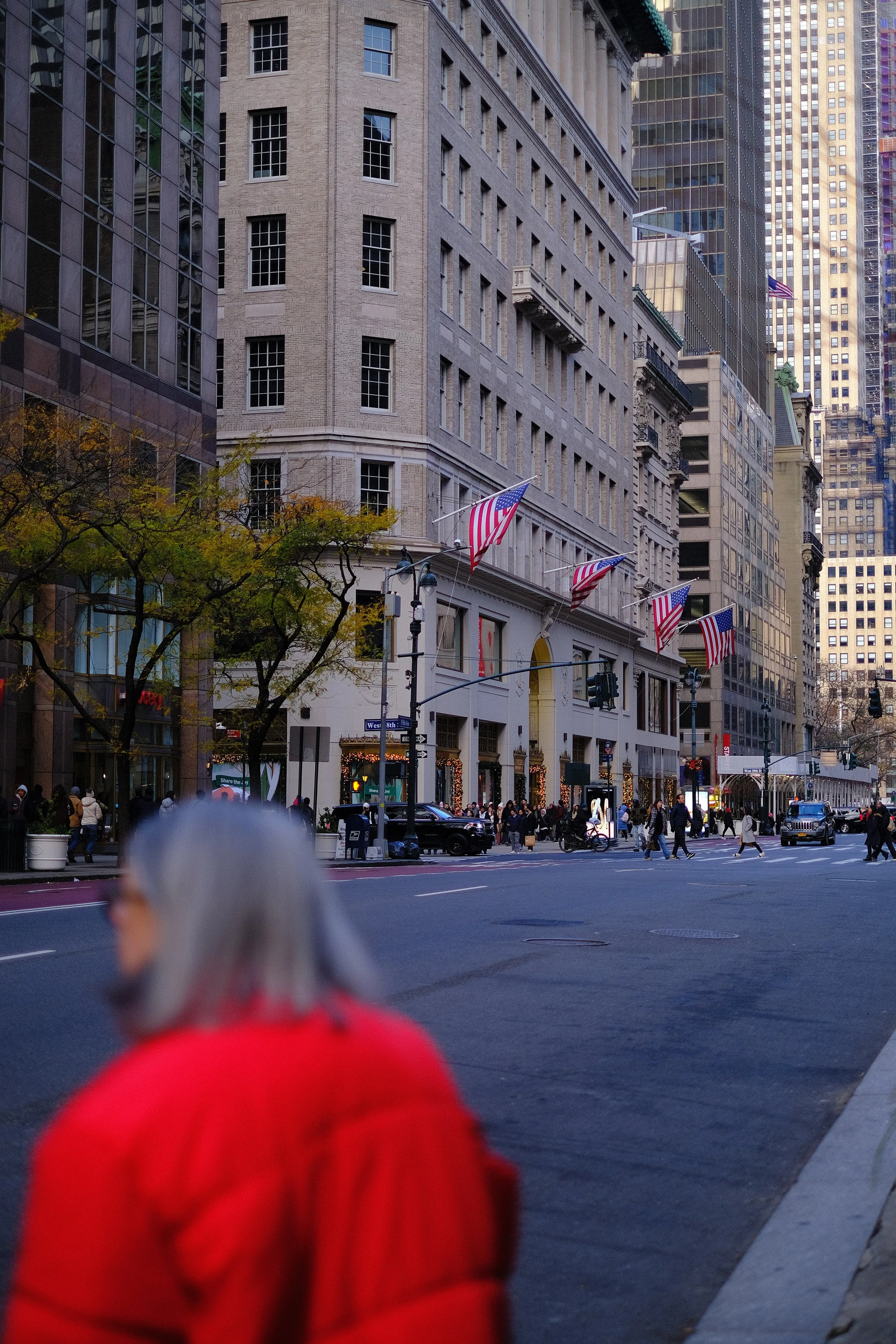 City street scene with pedestrians walking, buildings with American flags, and a person with gray hair and a red coat in the foreground.