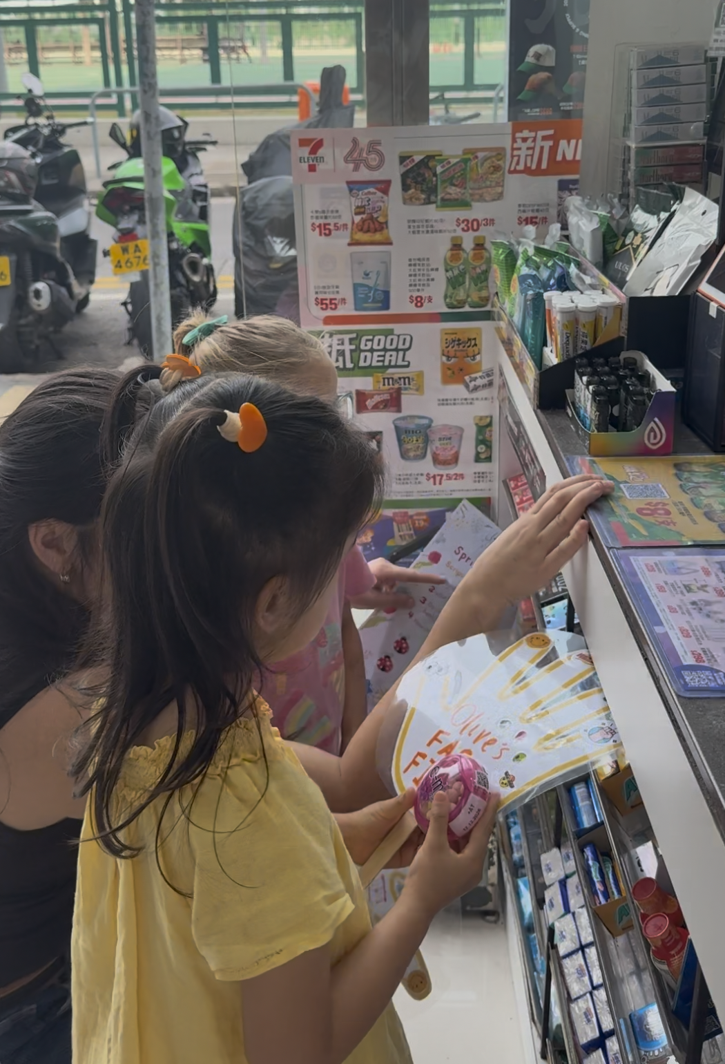 Three children and an adult at a convenience store counter looking at snack and drink options, with motorcycles visible outside the store window.