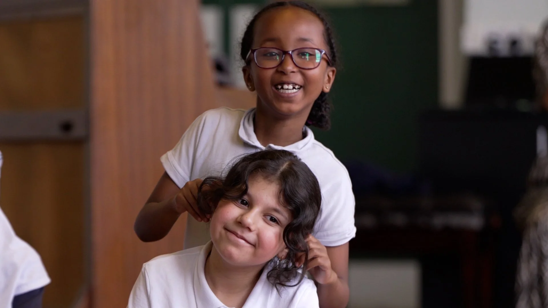 Two girls in white school uniforms smiling indoors. One girl with glasses is standing behind another girl who is sitting, and they are playing with her curly hair.