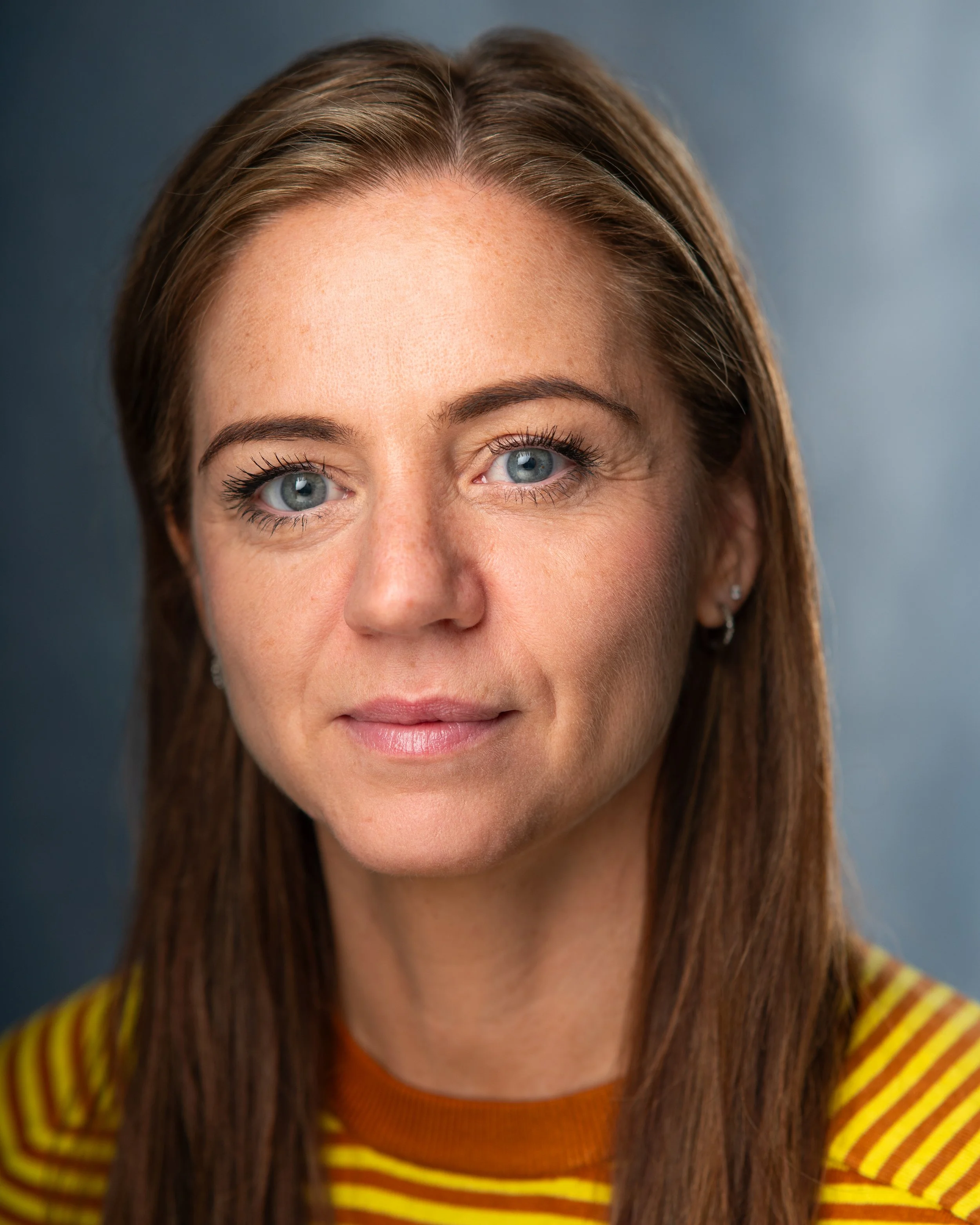 Close-up of a woman with light skin, blue eyes, brown hair, wearing a yellow and brown striped shirt, smiling slightly, with a neutral background.