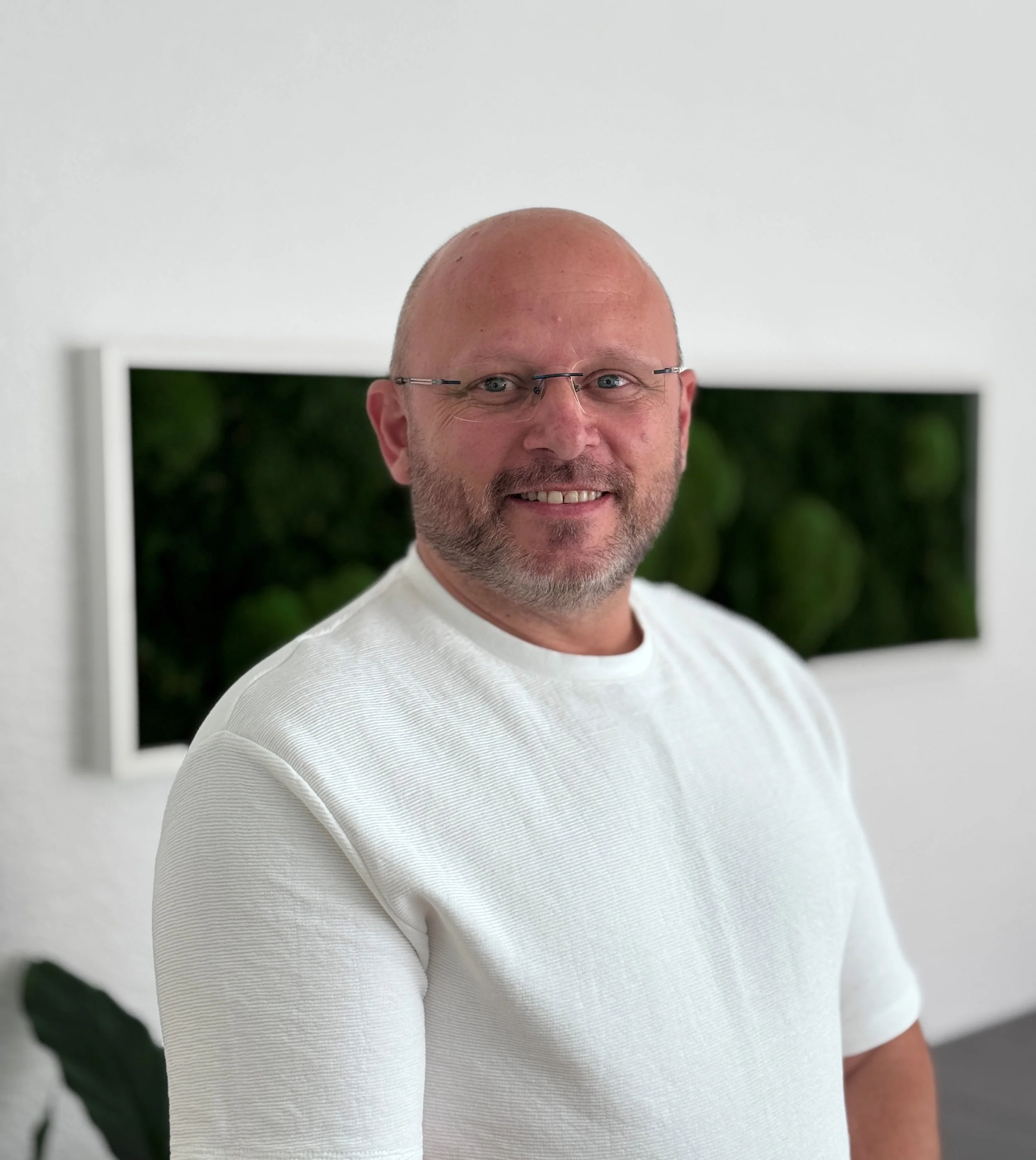 Porträt eines lachenden, glatzköpfigen Mannes mit Brille, der ein weißes T-Shirt trägt, vor einer weißen Wand mit dunklem Rahmen und grünen Pflanzen im Hintergrund.