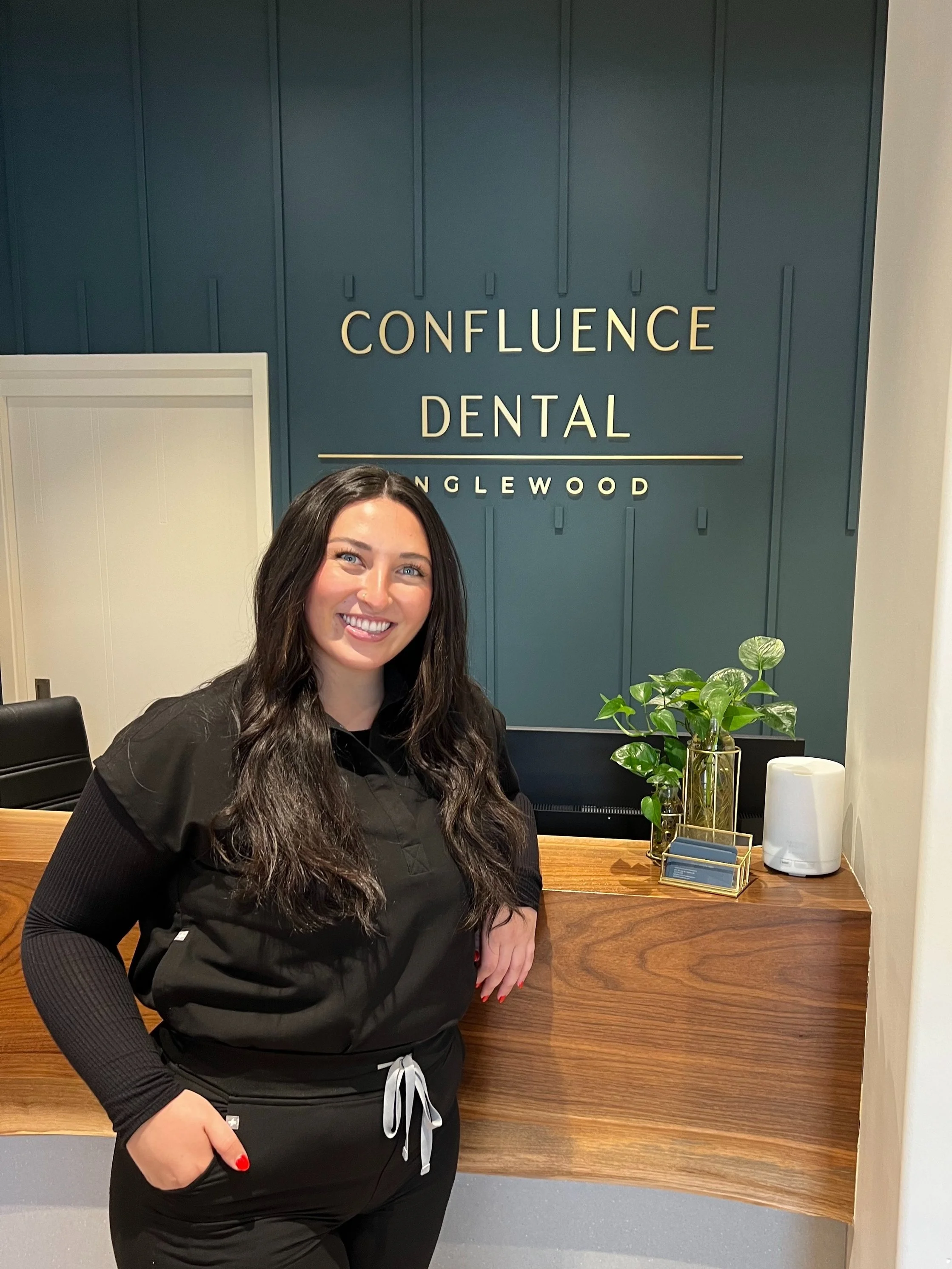 A woman standing in front of the reception desk at Confluence Dental, smiling. The reception area has a wooden counter, a potted plant, and dental supplies. The background features a blue wall with the dental office's sign.