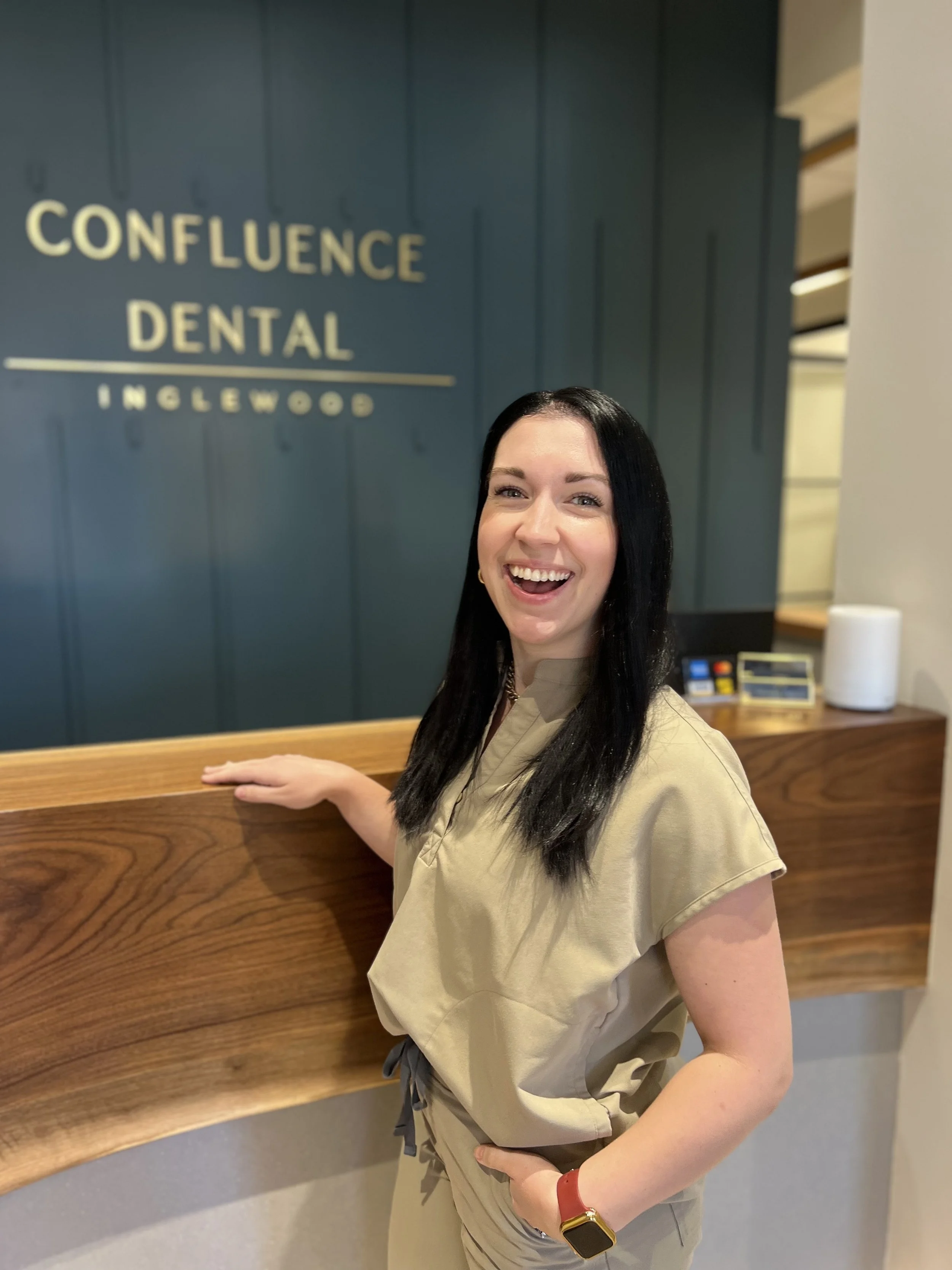 A woman with long black hair smiling at the camera, standing inside a dental office with a sign that reads 'Confluence Dental Inglewood' on the wall behind her.