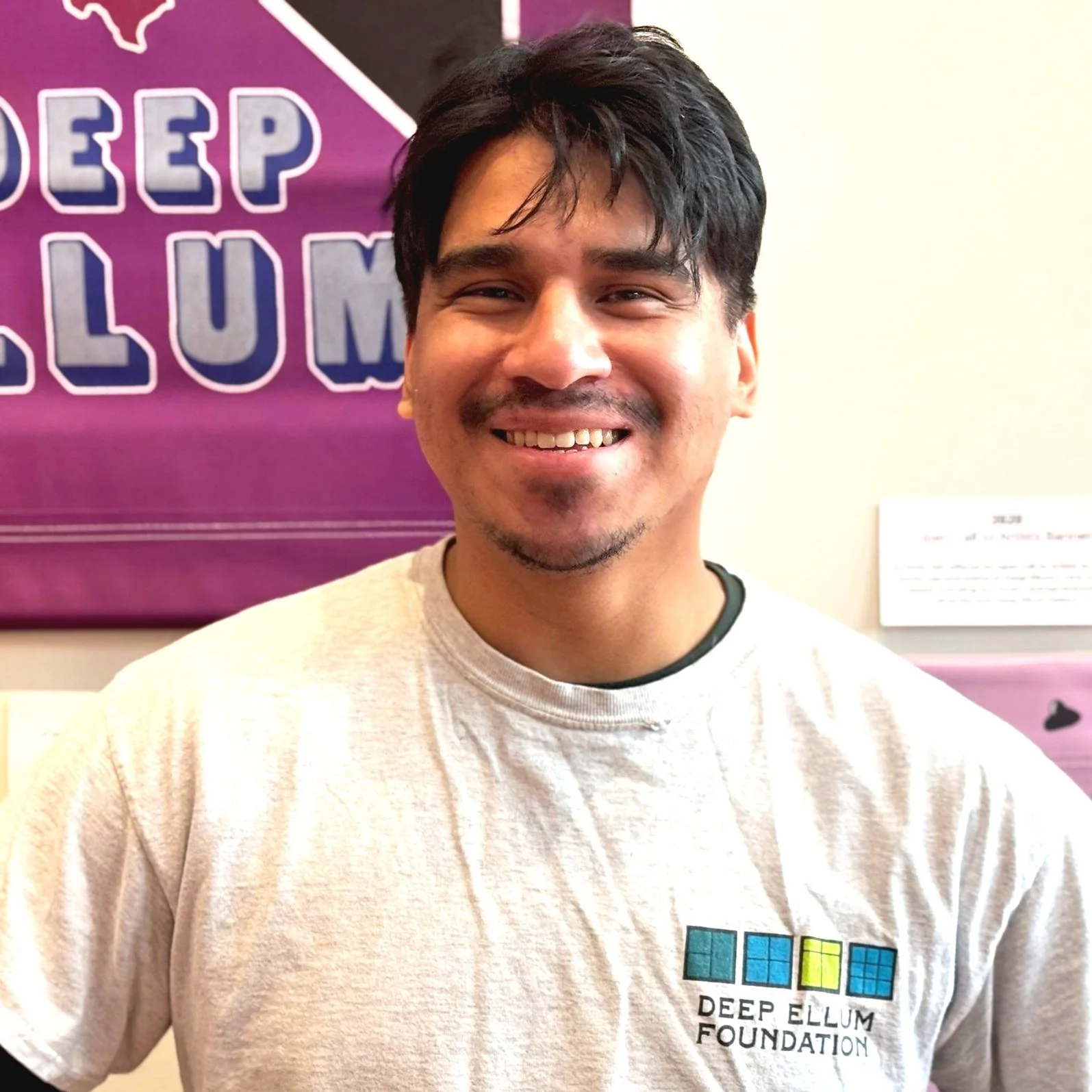 Smiling man with dark hair and a goatee wearing a white t-shirt with the Deep Ellum Foundation logo, standing indoors with a purple banner in the background.