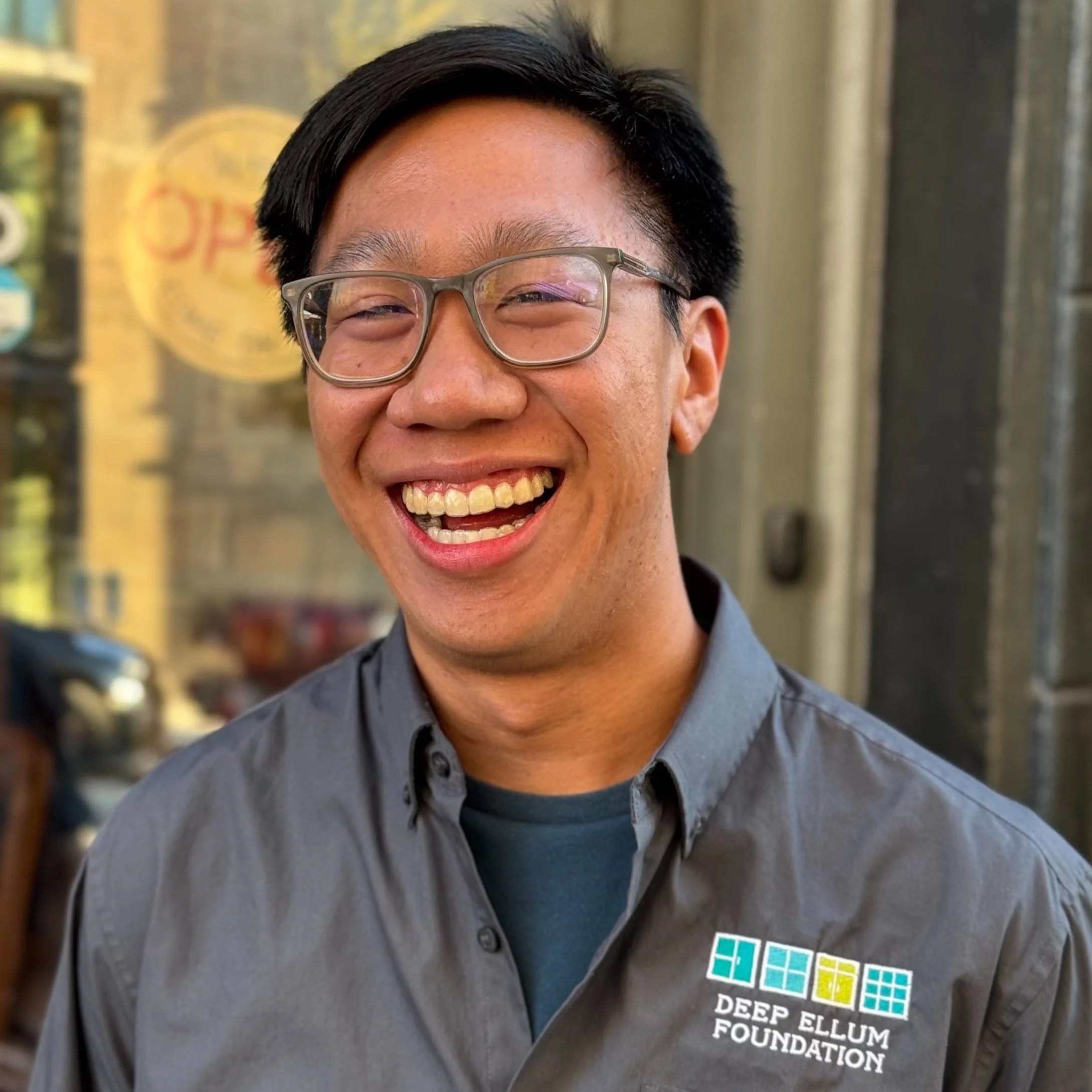 A smiling man wearing glasses and a gray shirt with the Deep Ellum Foundation logo, standing outdoors.