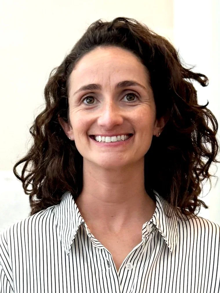 A woman with curly brown hair, smiling, wearing a white and black striped button-up shirt.