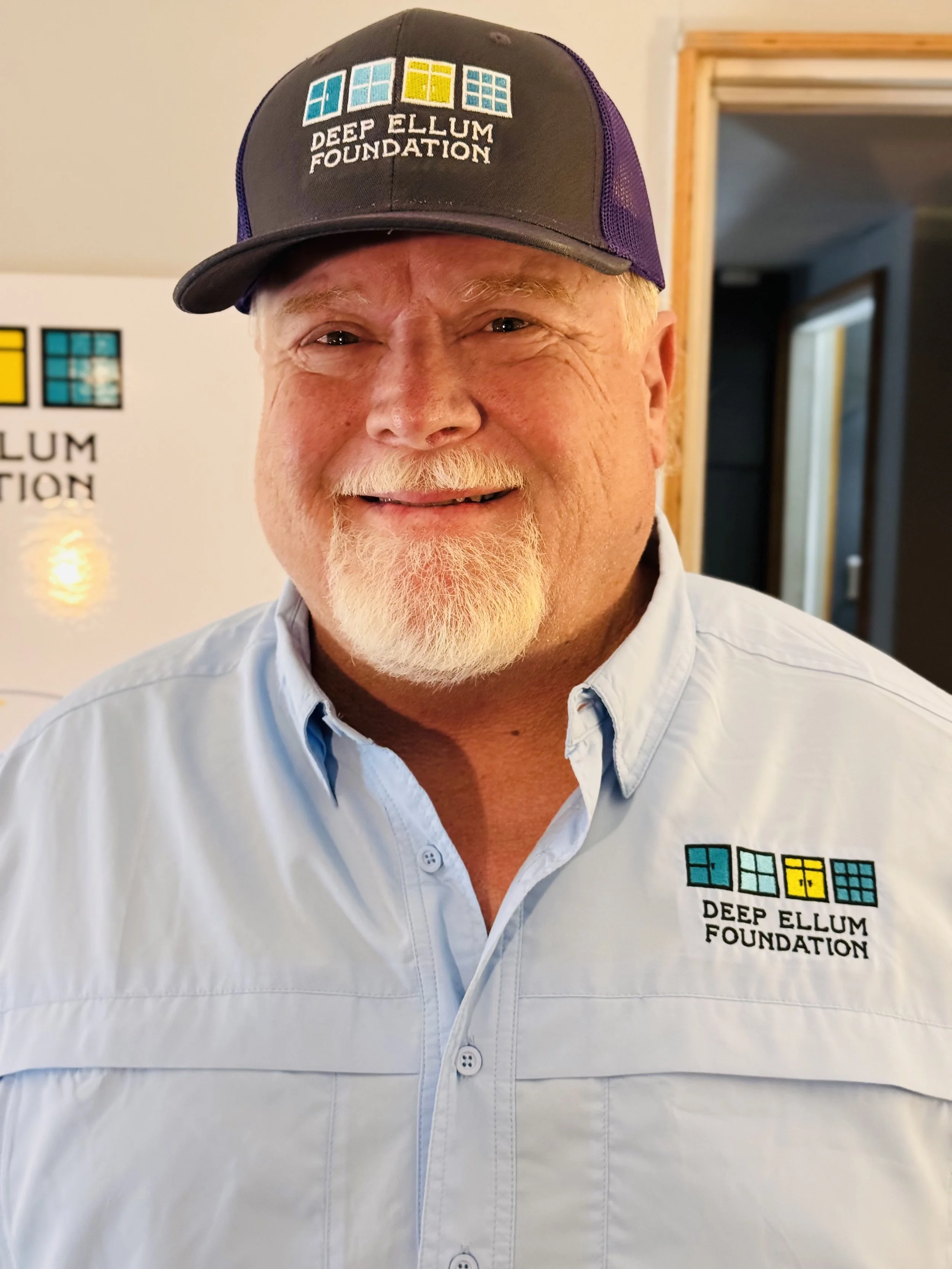 A smiling man wearing a light blue button-up shirt and a black and purple hat, both with the 'Deep Ellum Foundation' logo, standing indoors near a doorway with a white wall and a logo in the background.