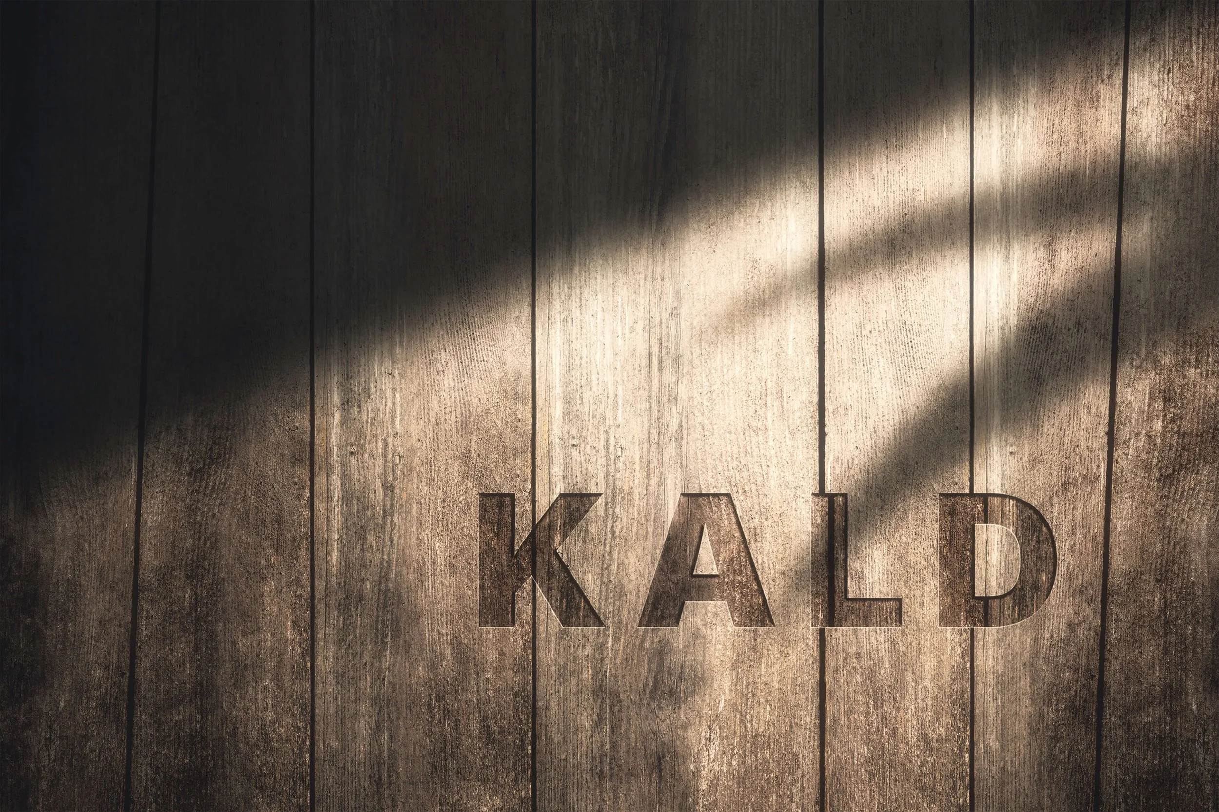 Close-up of a wooden surface with partially visible large metallic letters spelling 'KA' or 'KAD' and diagonal light beams crossing the surface.