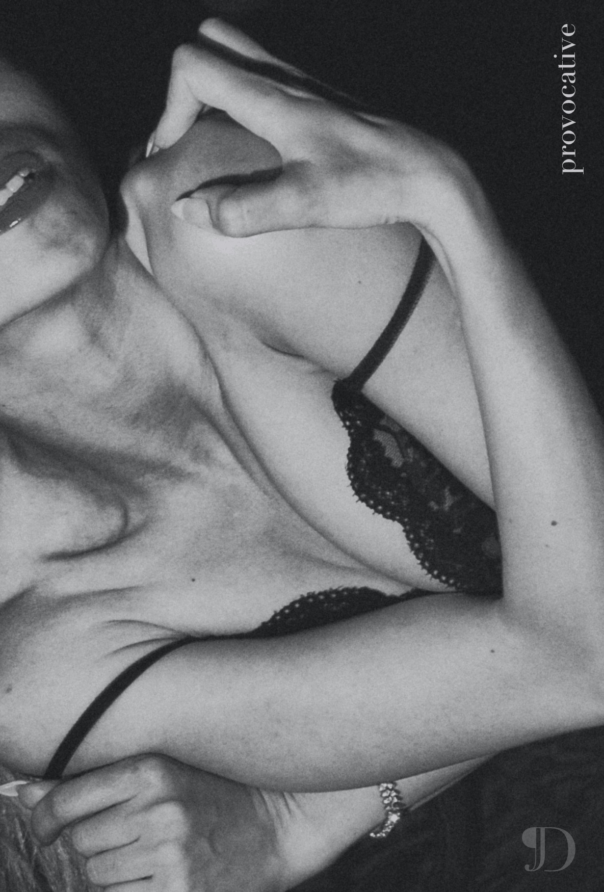Black and white photo of a woman lying down, wearing a black lace top, with her hand near her face, making an 'OK' gesture over her eye, smiling.