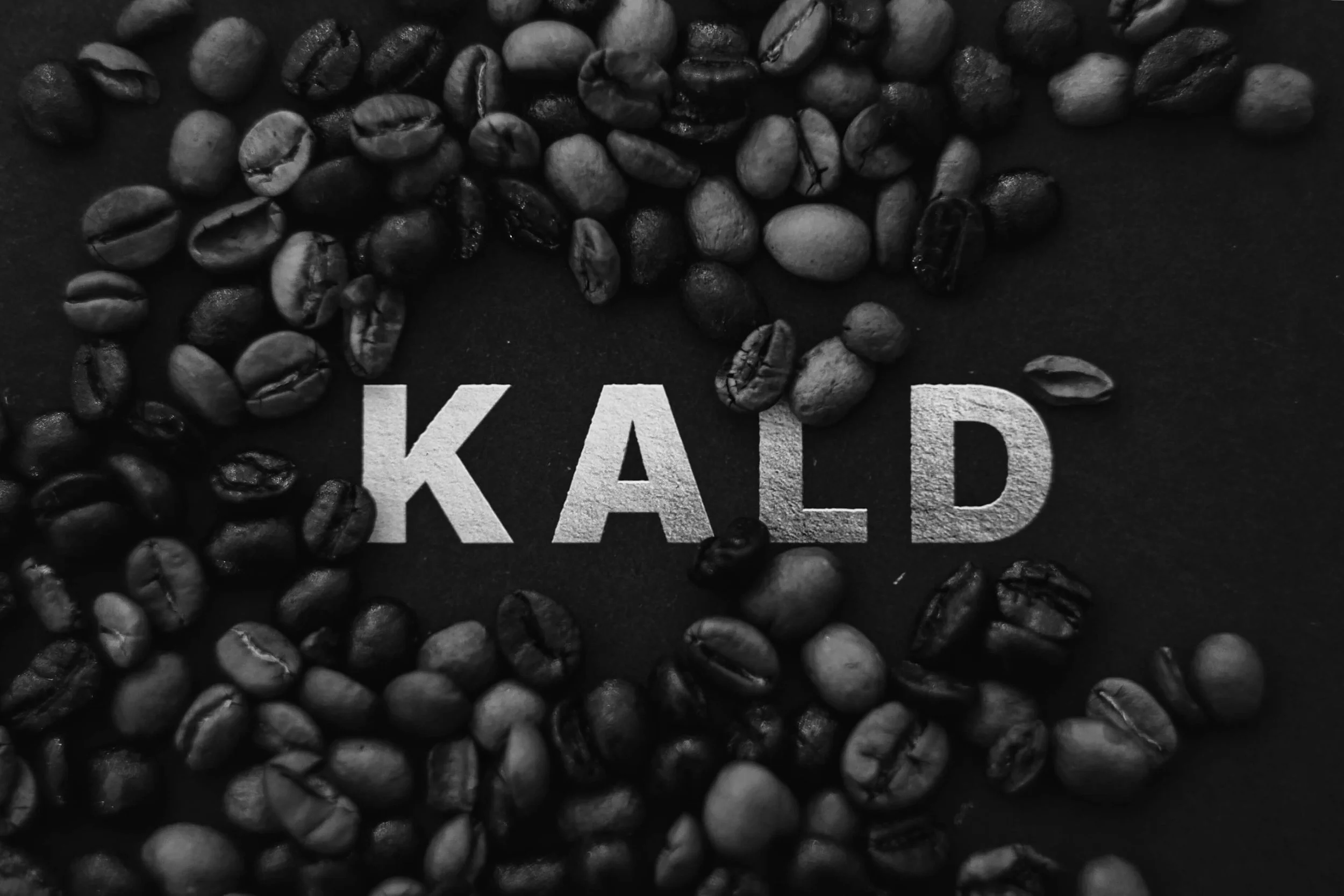 Close-up of roasted coffee beans scattered on a dark surface with partially visible white text reading 'KALD' underneath.