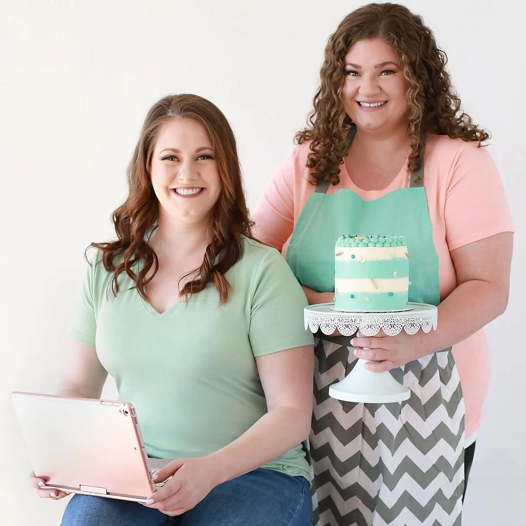 Two women smiling, one seated holding a tablet, the other standing behind holding a cake on a cake stand.