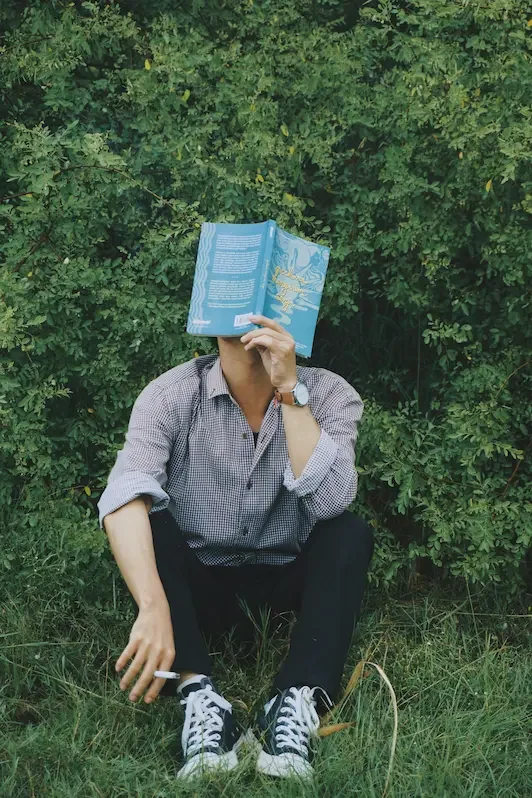 Person sitting on grass against dense greenery, holding a book over their face, suggesting withdrawal, introspection, and a desire to hide.