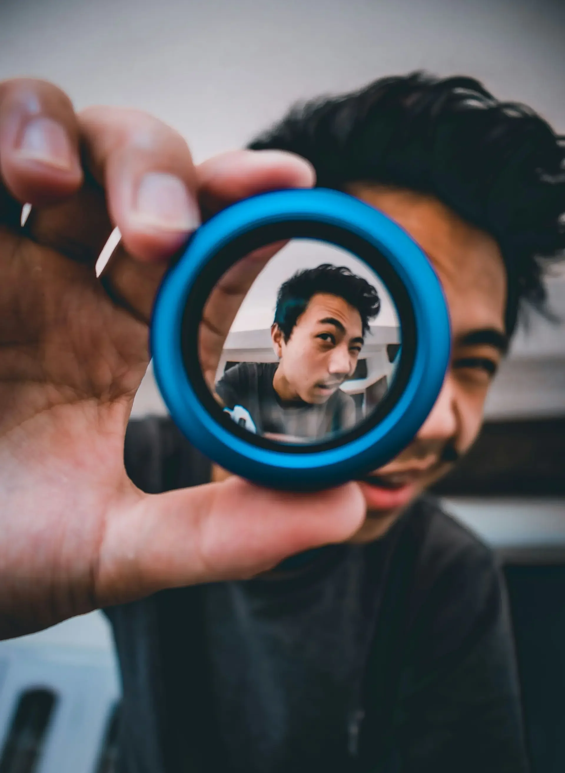 A creative portrait of a man holding a small, circular blue lens up to the camera. His face is blurred in the background, but his clear reflection is perfectly framed and in sharp focus within the center of the blue lens.