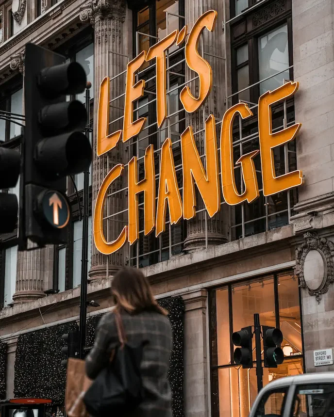 A close-up, low-angle shot of a large, bright orange neon sign on the side of a classic stone building that reads "LET’S CHANGE".