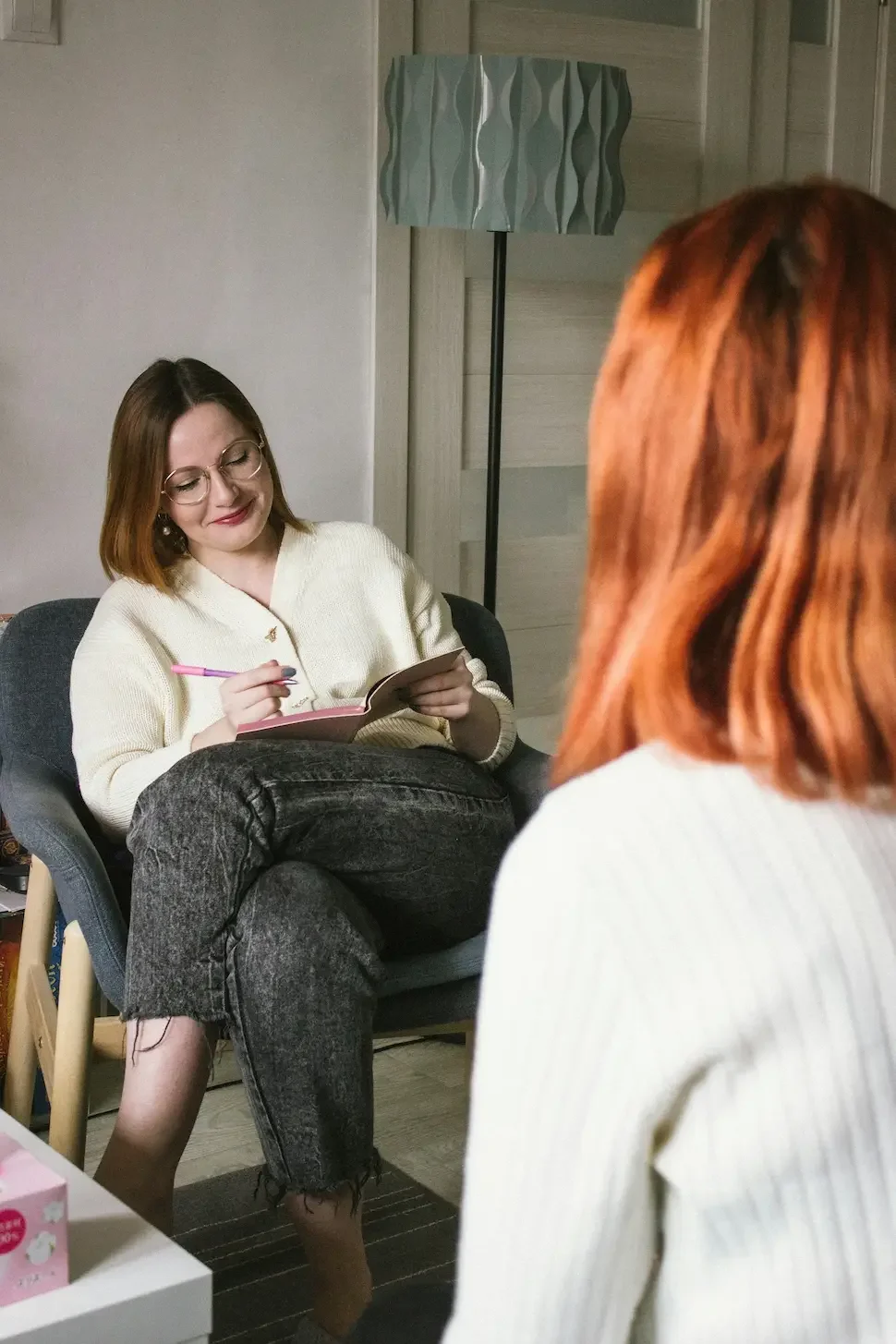 A therapist, wearing glasses and a cream-colored cardigan, is taking notes during a session with a client, whose red hair is visible from the back.
