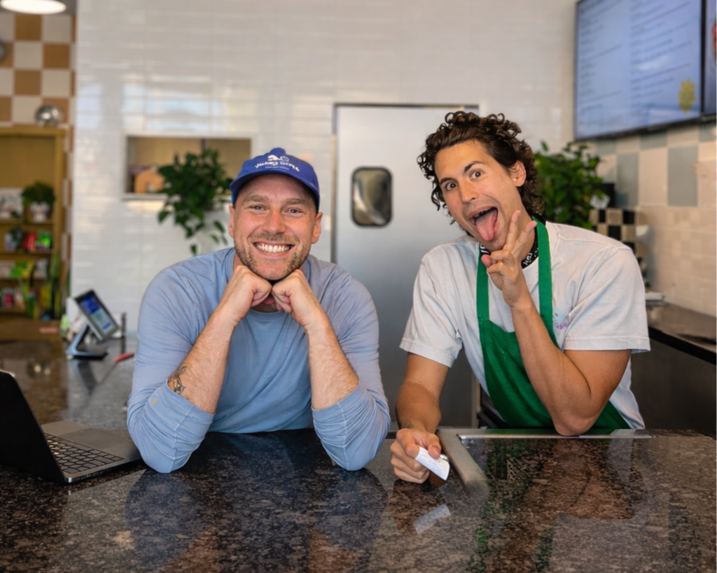Two men are sitting at a counter in a restaurant, smiling at the camera. One man wears a blue cap and long-sleeve shirt, and the other wears a white shirt with green apron and is making a playful face with his tongue out.