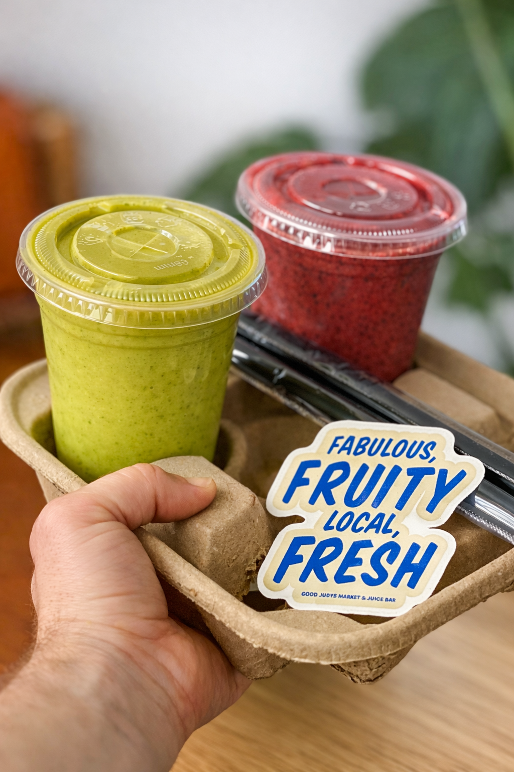 A hand holding a cardboard tray with two smoothies, one green and one red, and a sticker that reads "Fabulous, Fruity, Local, Fresh". There are black straws and a small brown paper package on the tray.