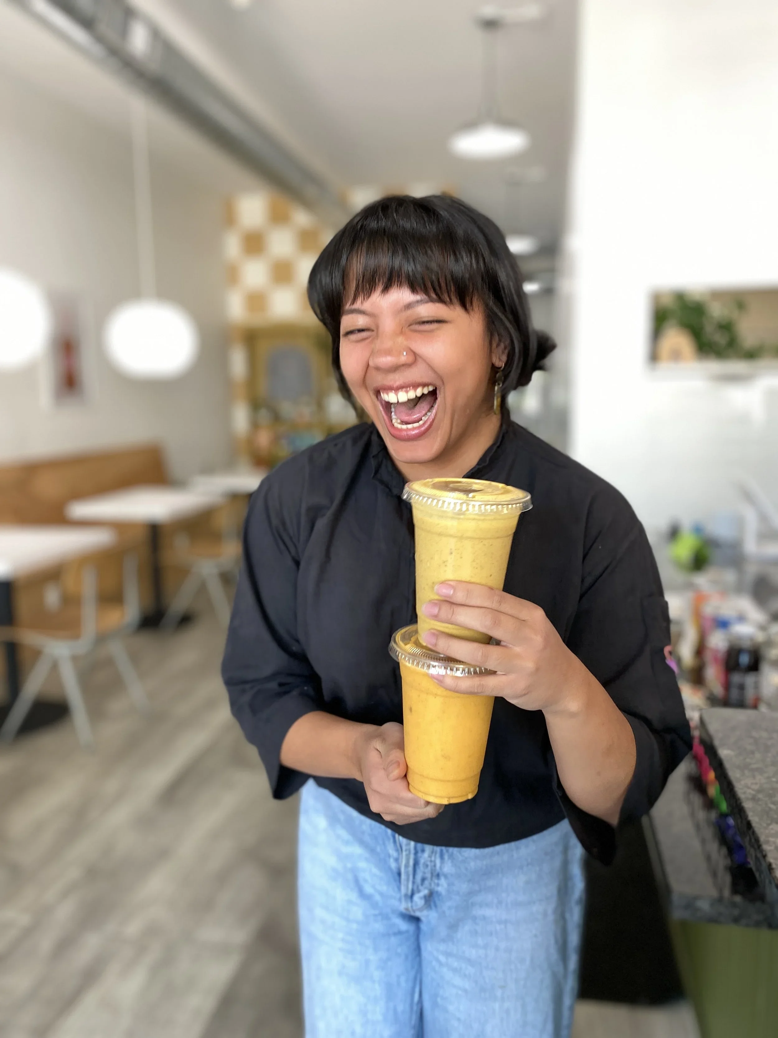 Person with short dark hair laughing and holding two yellow smoothies in a cafe.