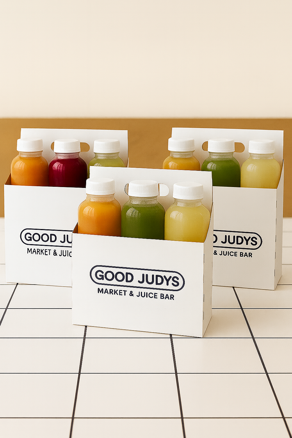 Three white boxes labeled 'Good Judys Market & Juice Bar' containing bottles of colorful fruit and vegetable juices on a tiled surface.