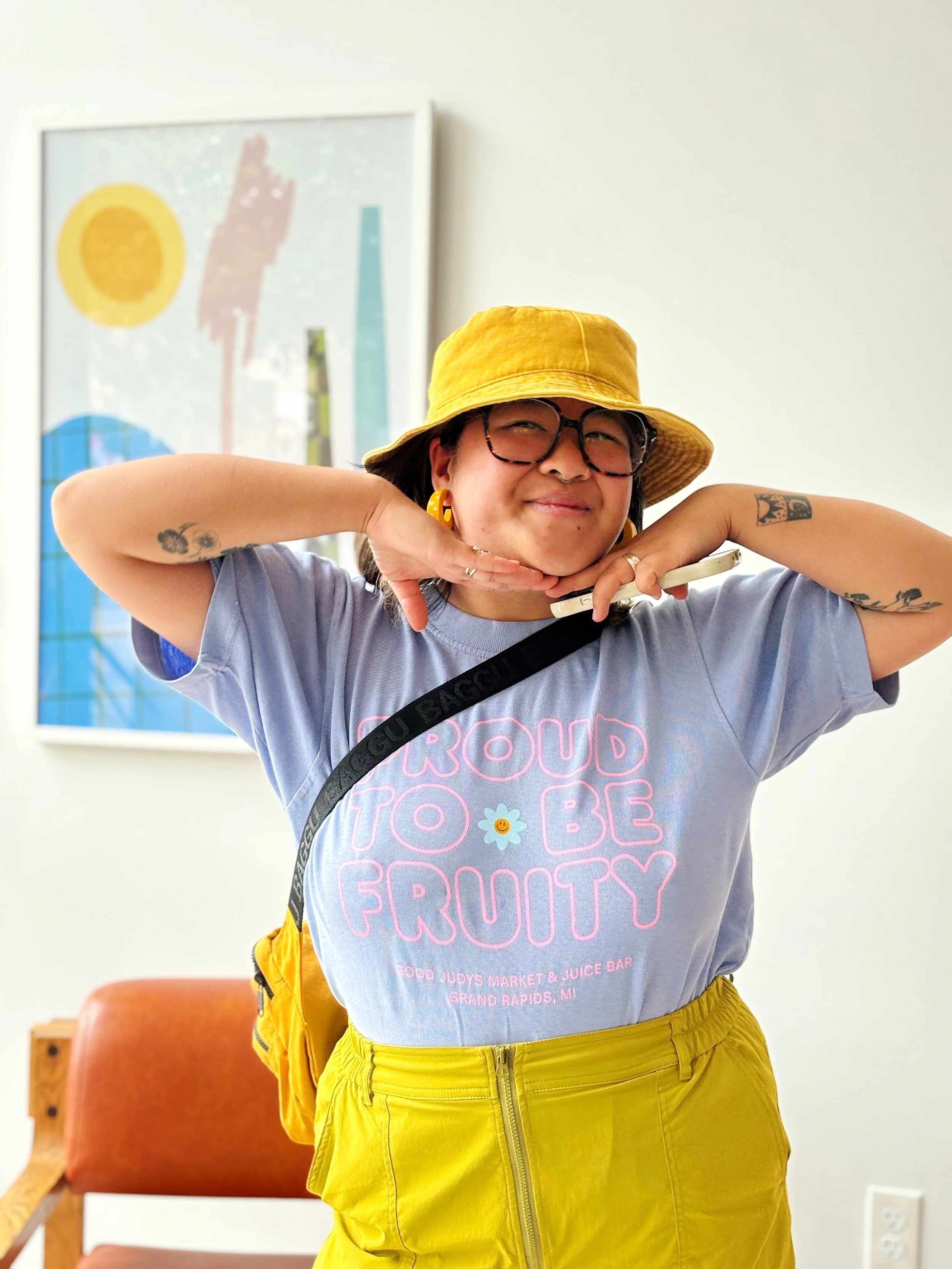 Person with a yellow hat and yellow pants, wearing glasses and a graphic T-shirt, smiling with hands under chin, standing indoors with artwork on the wall behind.