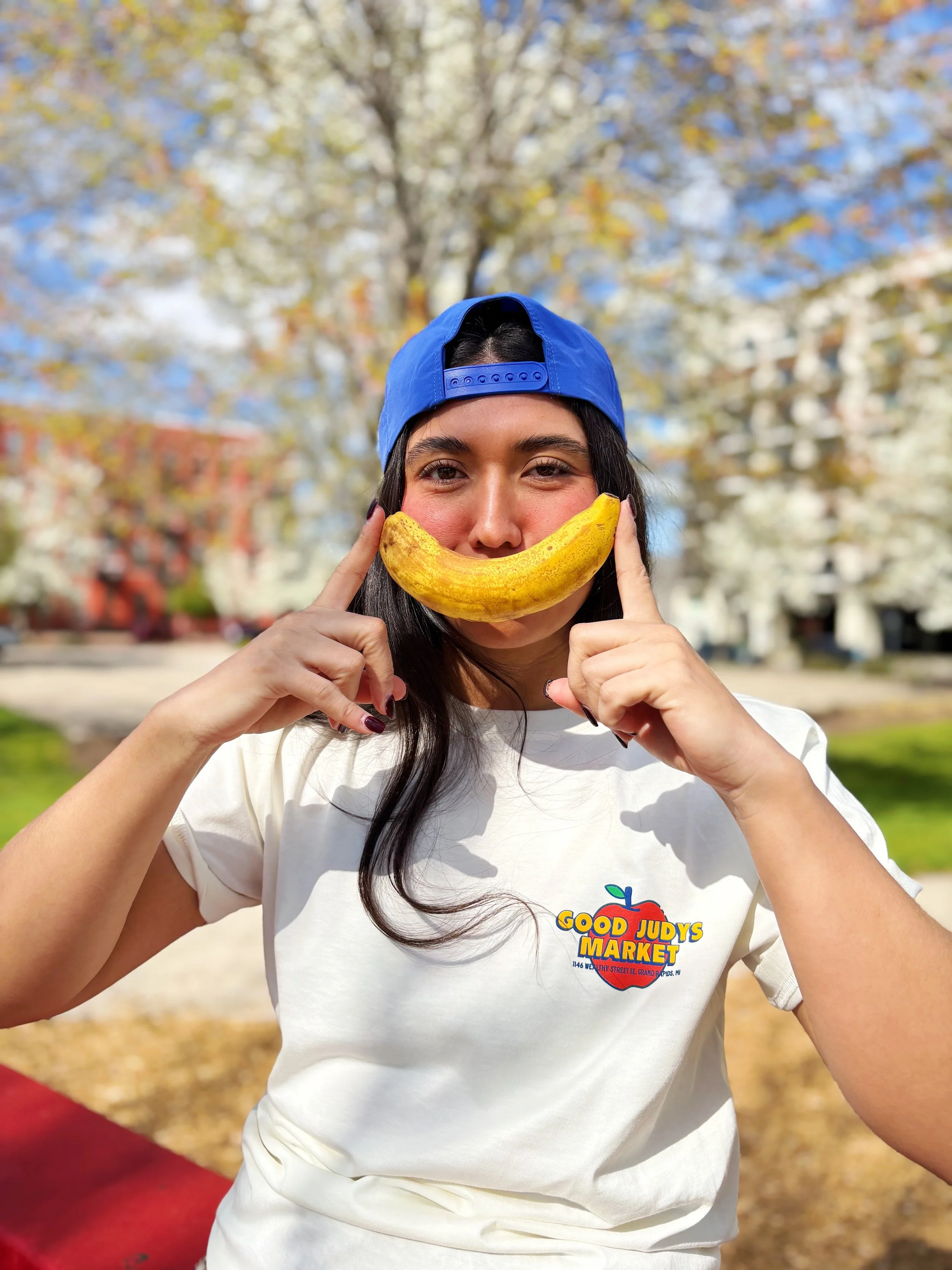 Smiling girl in Grand Rapids wearing a Good Judy’s Market T-shirt and holding a banana at a queer-owned smoothie and juice bar