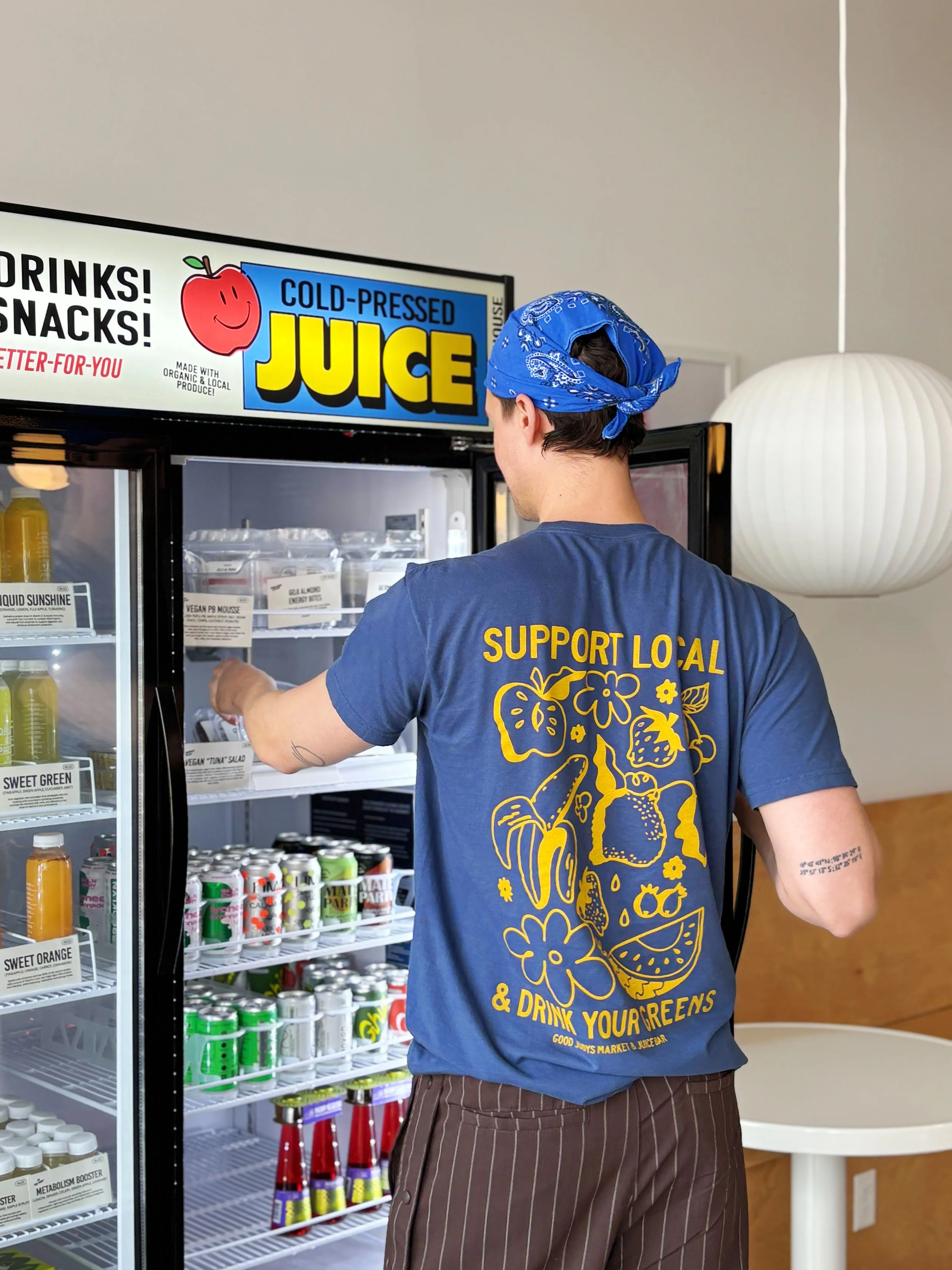 A person wearing a blue shirt with a yellow graphic and text supporting local efforts, selecting a drink from a fridge labeled 'Cold-Pressed Juice'. The person has a blue bandana on their head and is viewing various canned and bottled beverages.