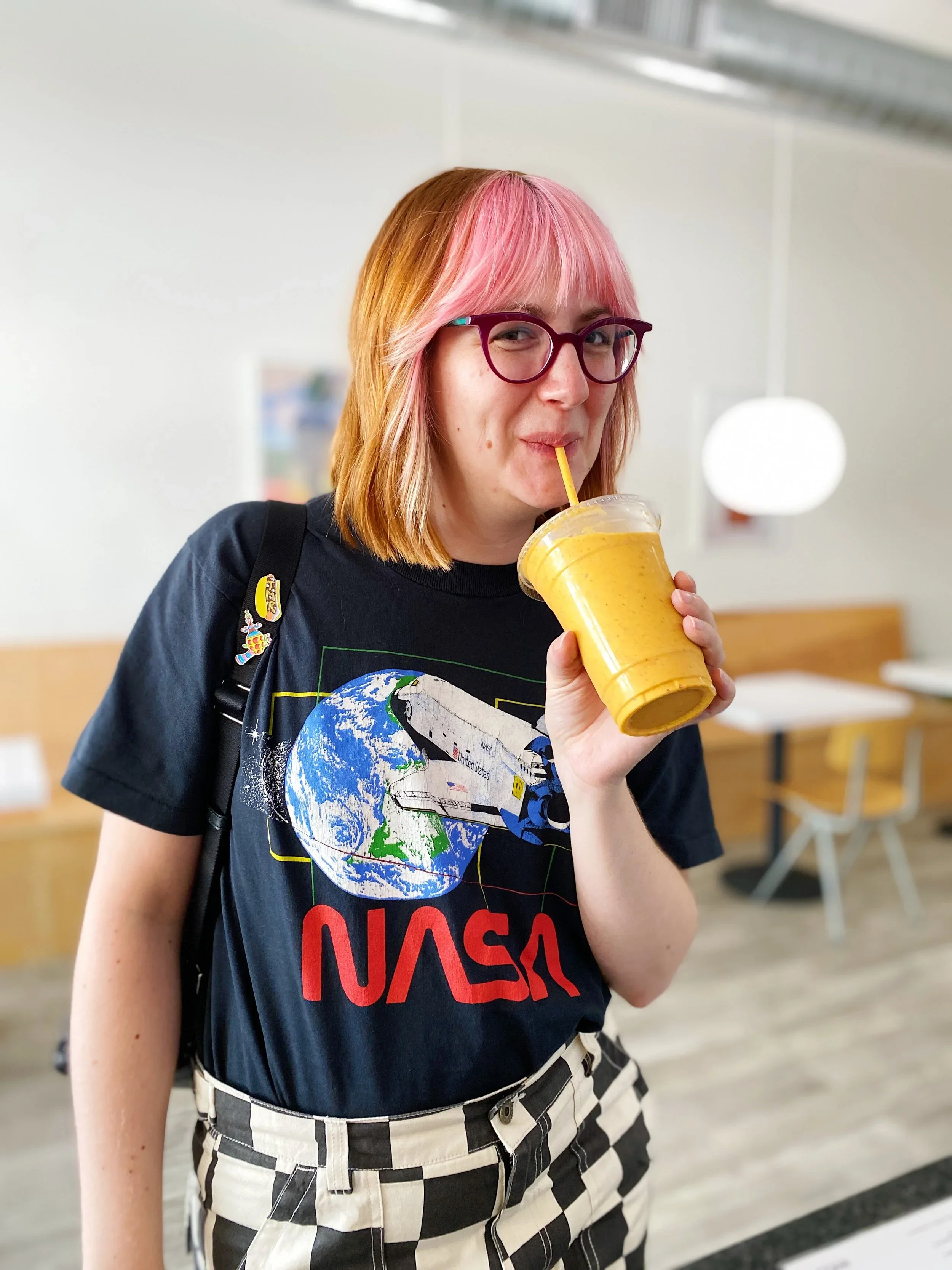A young woman with pink and blonde hair, wearing glasses, a NASA t-shirt, and checkered pants, drinks a yellow smoothie through a straw in an indoor setting.