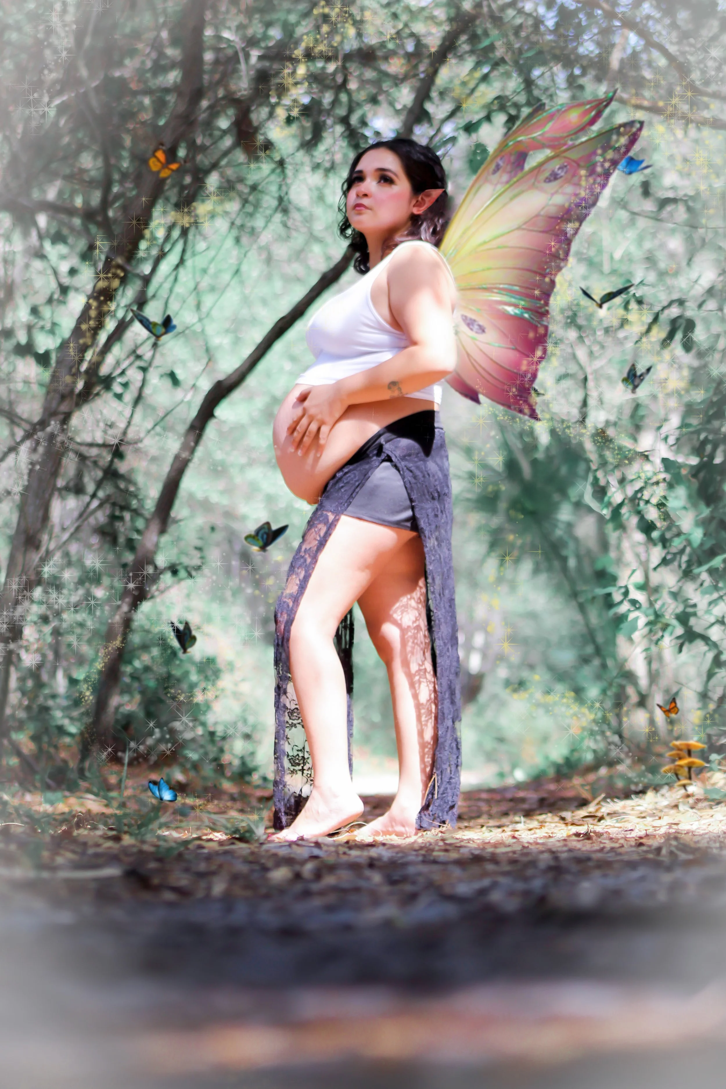 A woman with fairy wings standing outdoors in a wooded area, surrounded by butterflies and glowing sparkles.