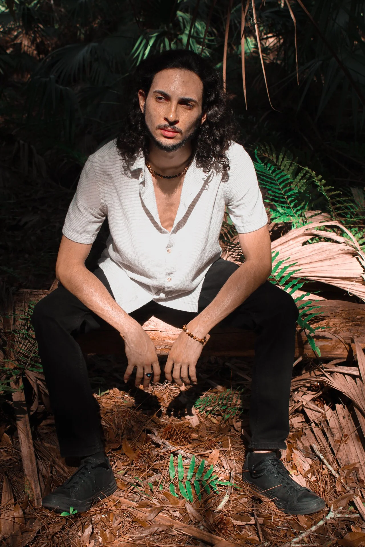 A man with long dark hair sitting on a log in a forest, wearing a light-colored shirt, black pants, and black shoes. He is looking pensively into the distance with sunlight highlighting his face and surroundings.